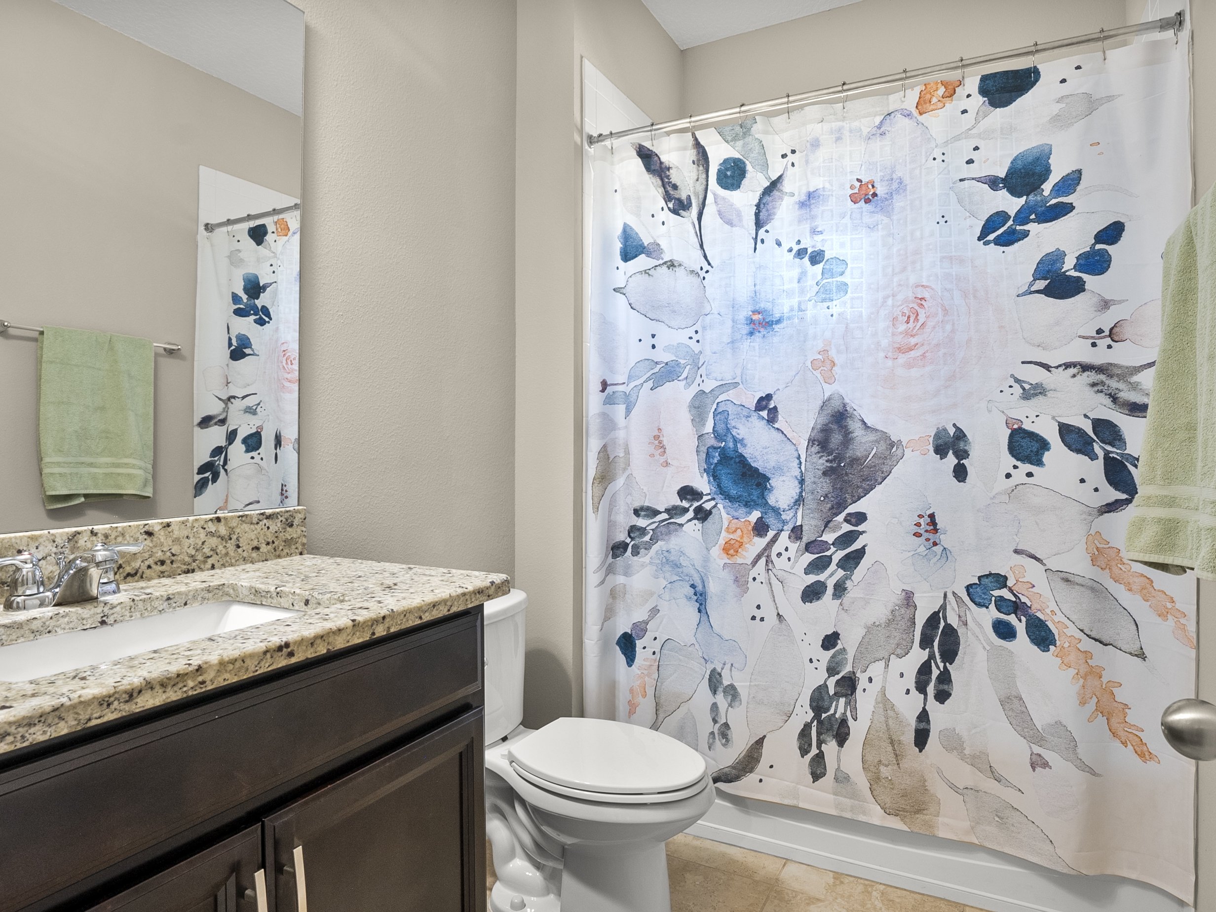 Bathroom with floral shower curtain, beige walls, tiled floor, granite countertop sink, and green towels.
