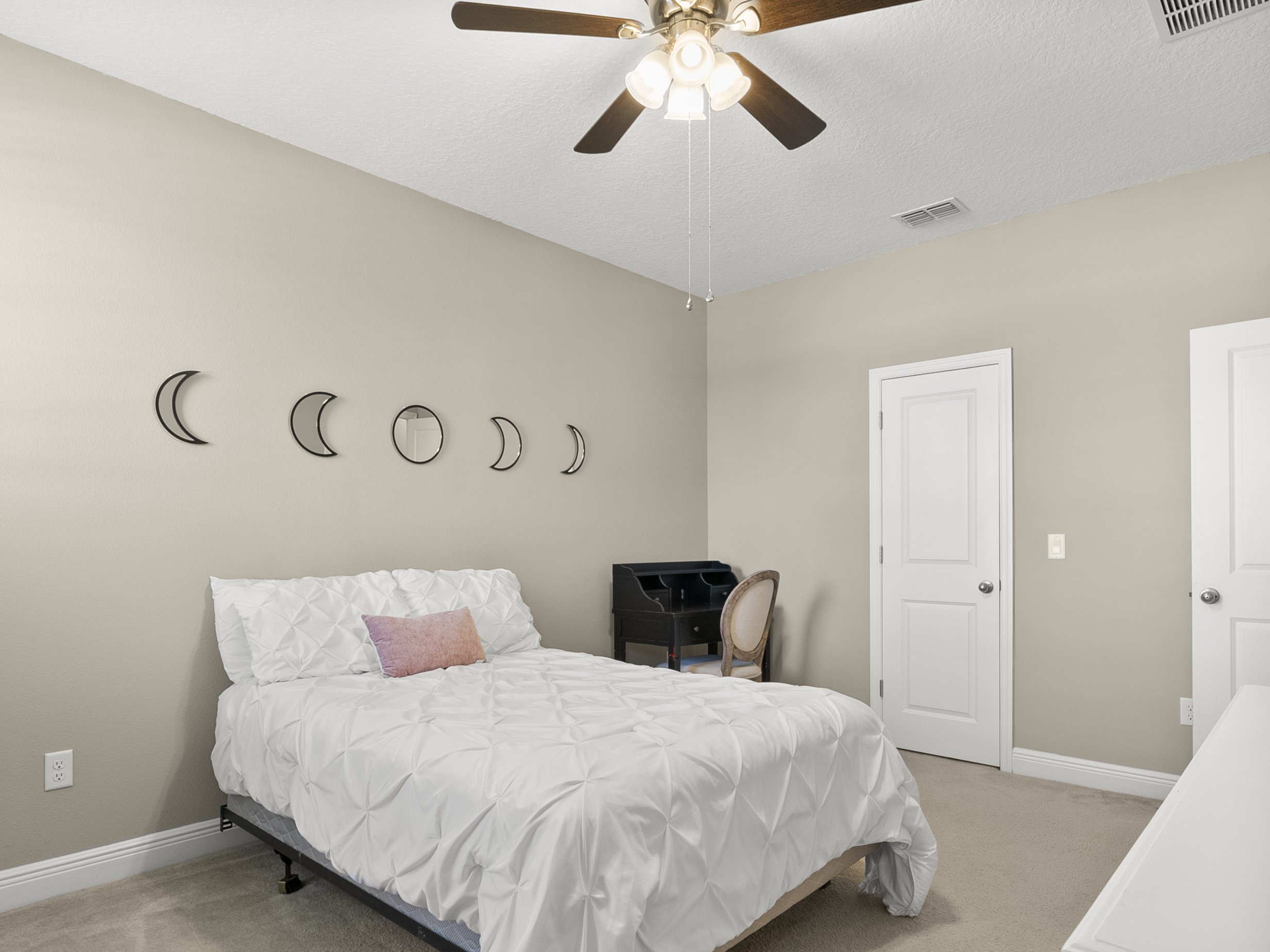 Bedroom with beige walls, a ceiling fan, a neatly made bed with white bedding, a pink pillow, moon phase wall art, a black desk, a chair, and two closed white doors.