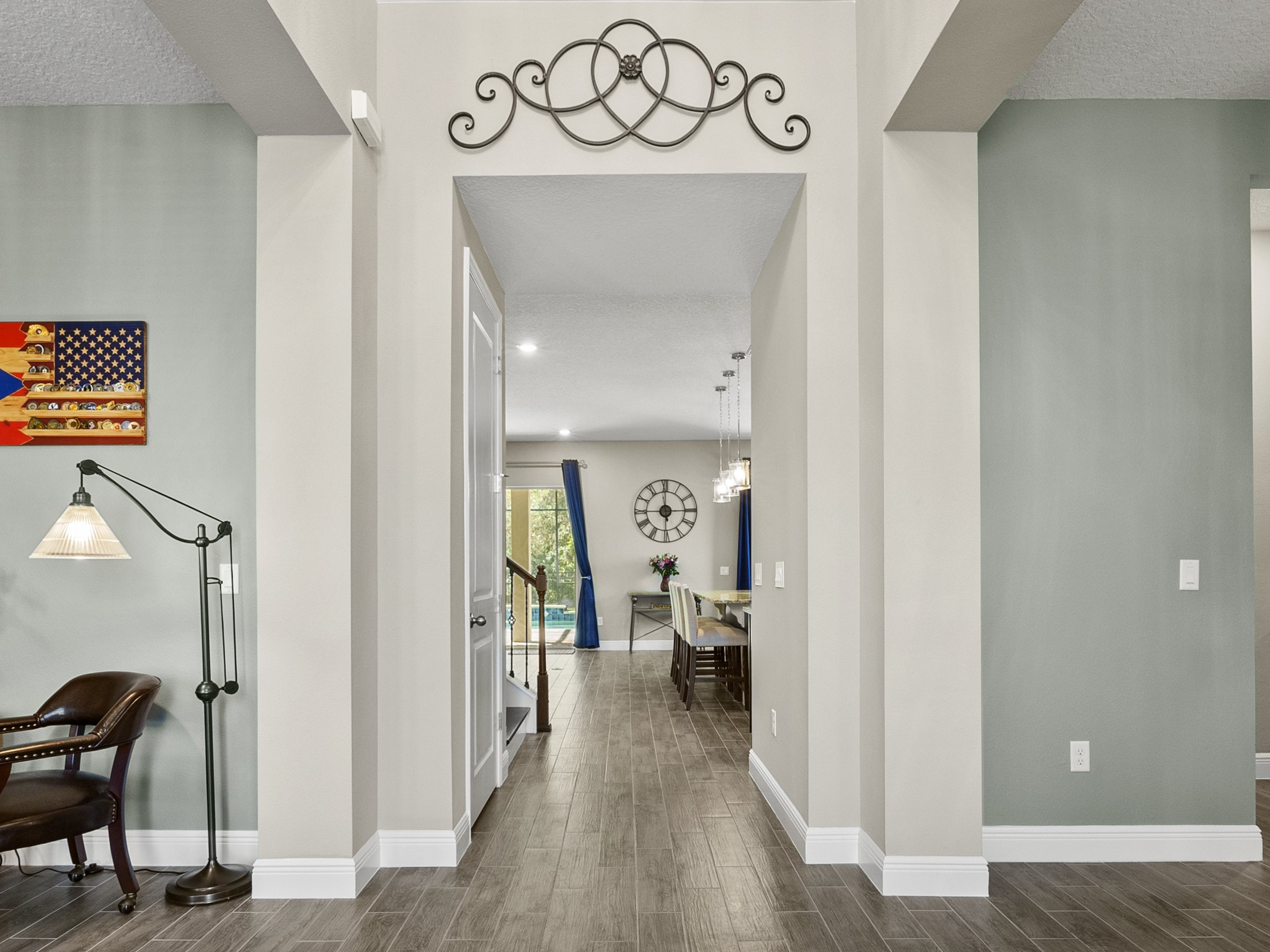 Interior hallway leading to a dining area with large wall clock, modern chandelier, and blue curtains. Decorative wall art and lamp are visible on the left side.