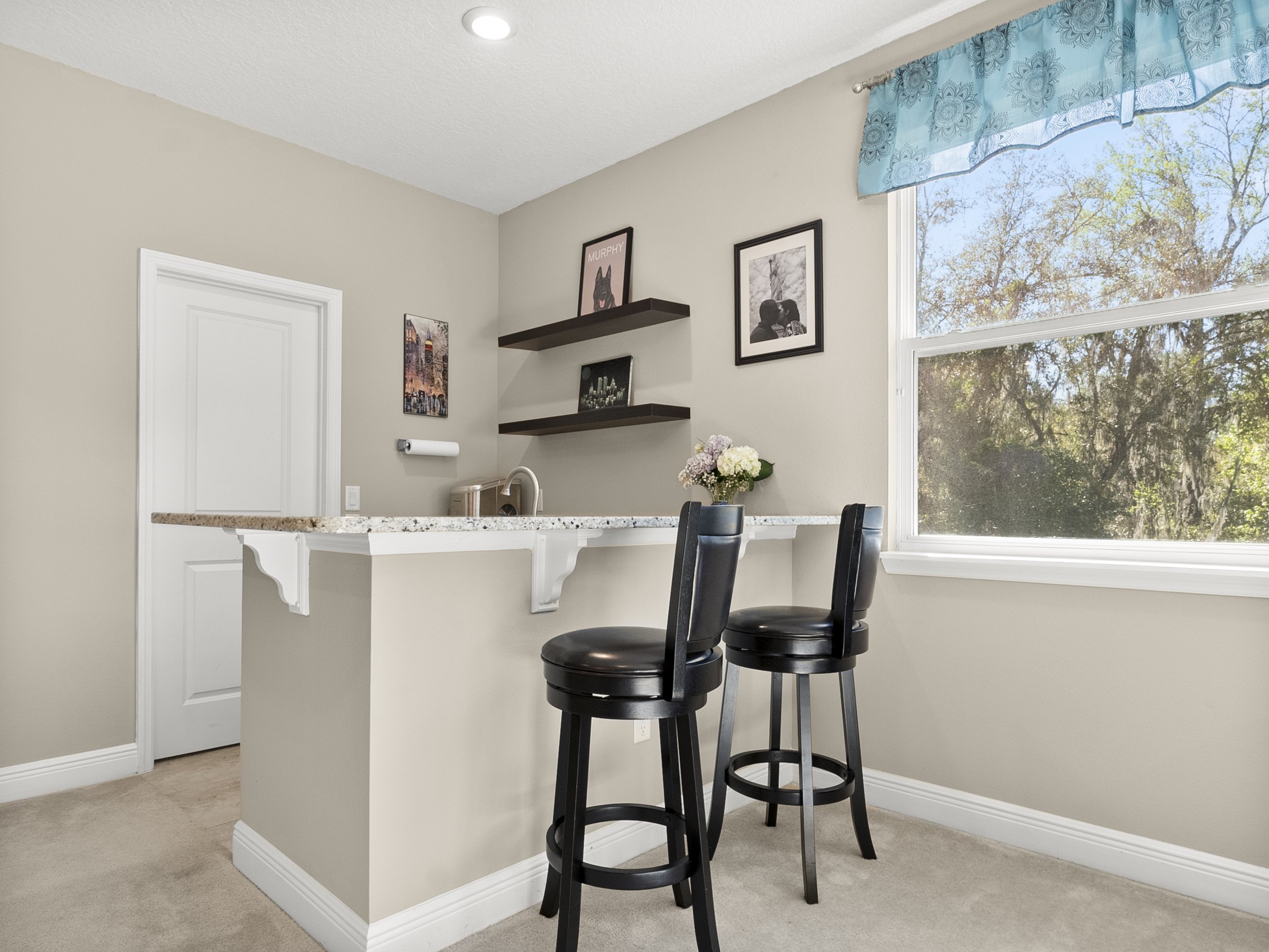 Small home bar with two black stools, granite countertop, wall shelves with decor, and a window with blue curtains.