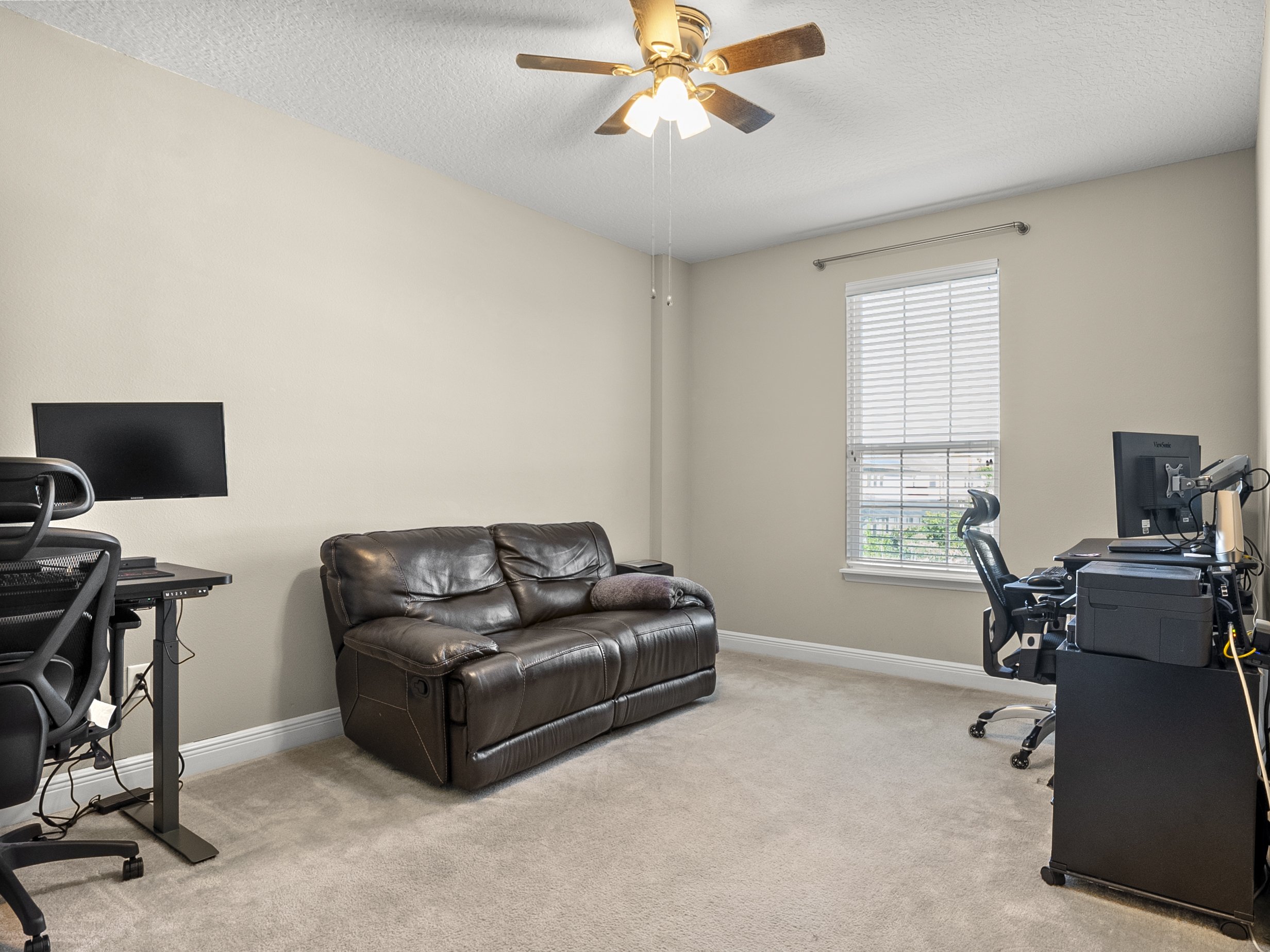 Home office with two desks and a brown leather couch. Each desk has a computer monitor and ergonomic chair. A ceiling fan is above, and a window with blinds allows natural light into the room. The floor is carpeted.