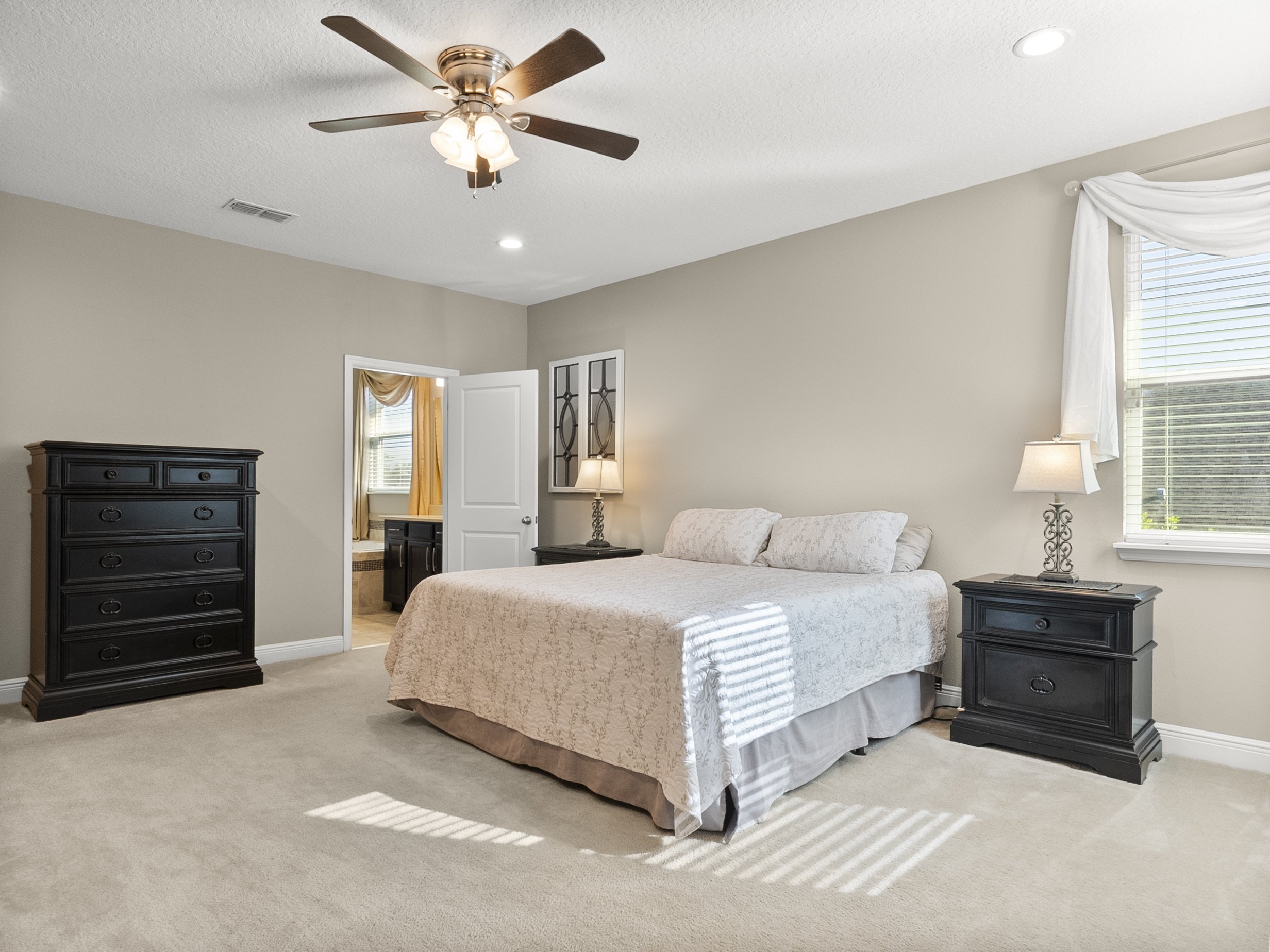 Bedroom interior with neutral walls, a ceiling fan, a bed with light-colored bedding, two matching black nightstands with lamps, a black dresser, large window with blinds, and a doorway to a bathroom.