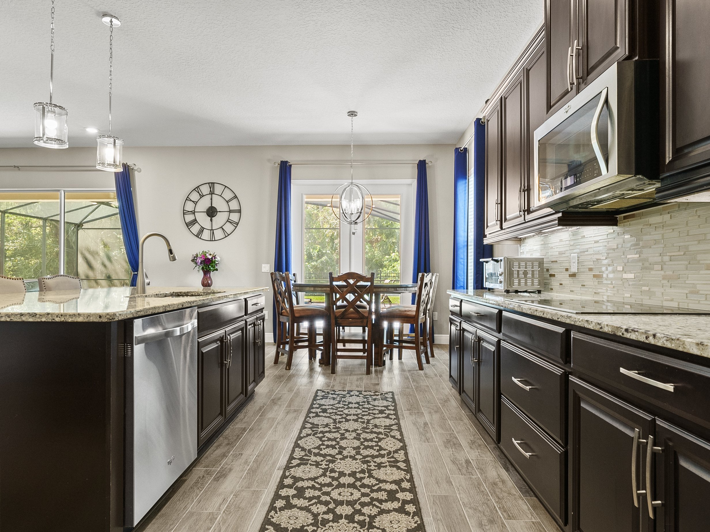 Modern kitchen and dining area with dark wood cabinets, stainless steel appliances, and granite countertops. The space is decorated with blue curtains, a large wall clock, pendant lights, and a central dining table with chairs. A floral arrangement a