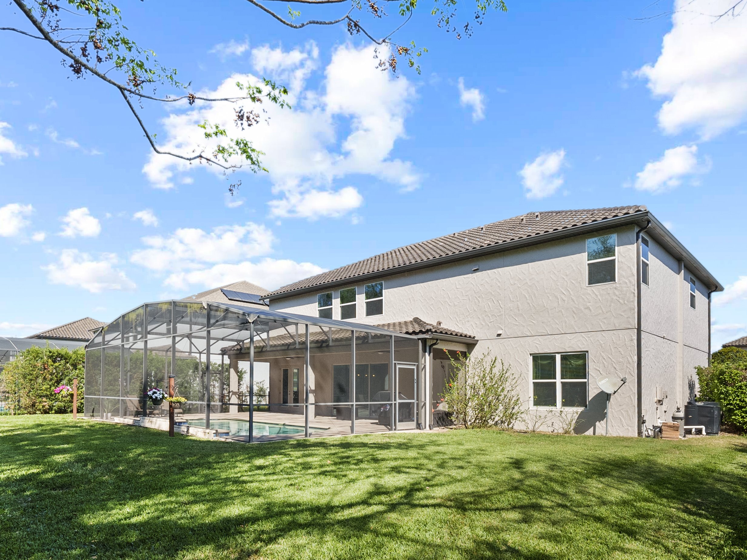 Backyard view of a two-story house with a screened-in porch and pool, surrounded by a well-maintained lawn, under a clear blue sky with scattered clouds.