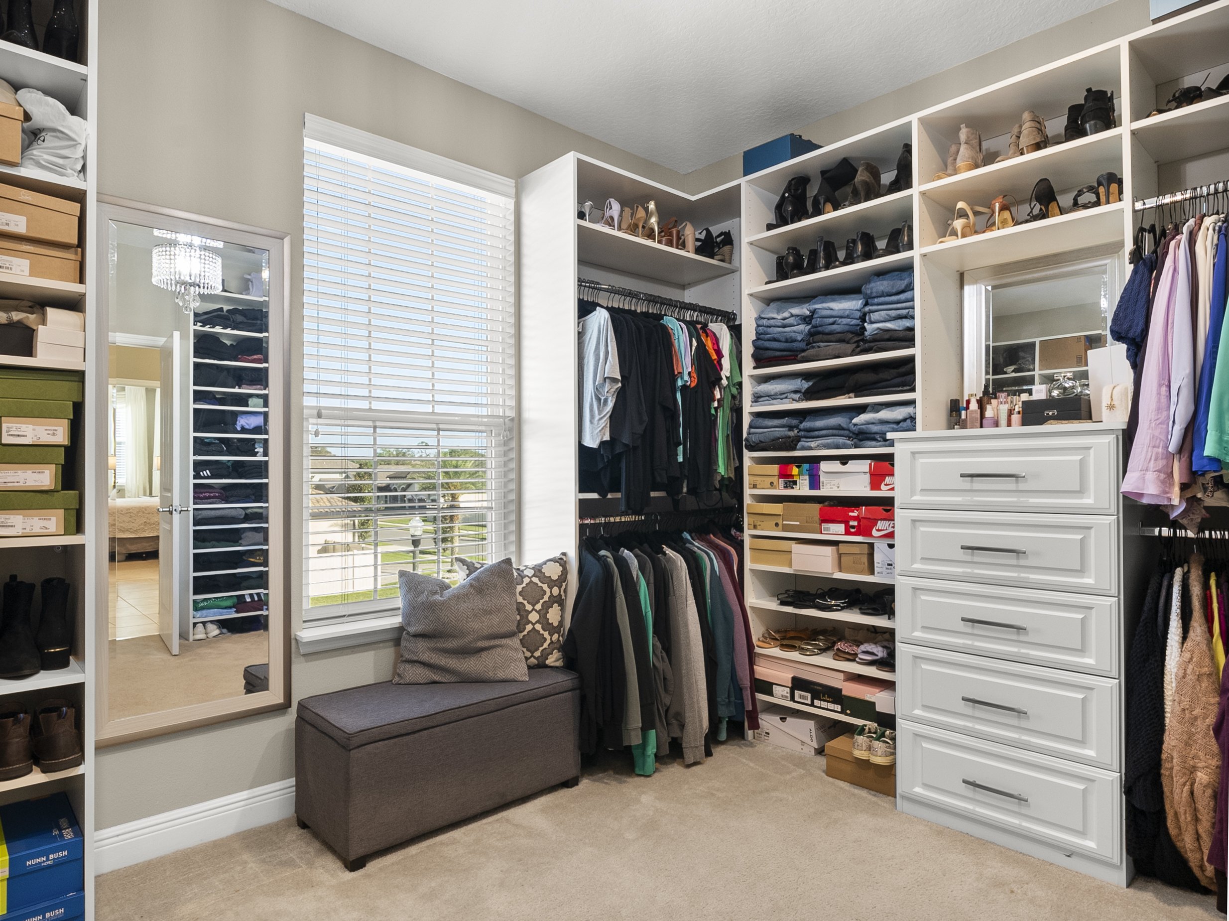 Walk-in closet with shelves and drawers filled with clothes, shoes, and accessories, including jeans, shirts, jackets, and various footwear. A bench is positioned below a window, and a full-length mirror is mounted on the wall.