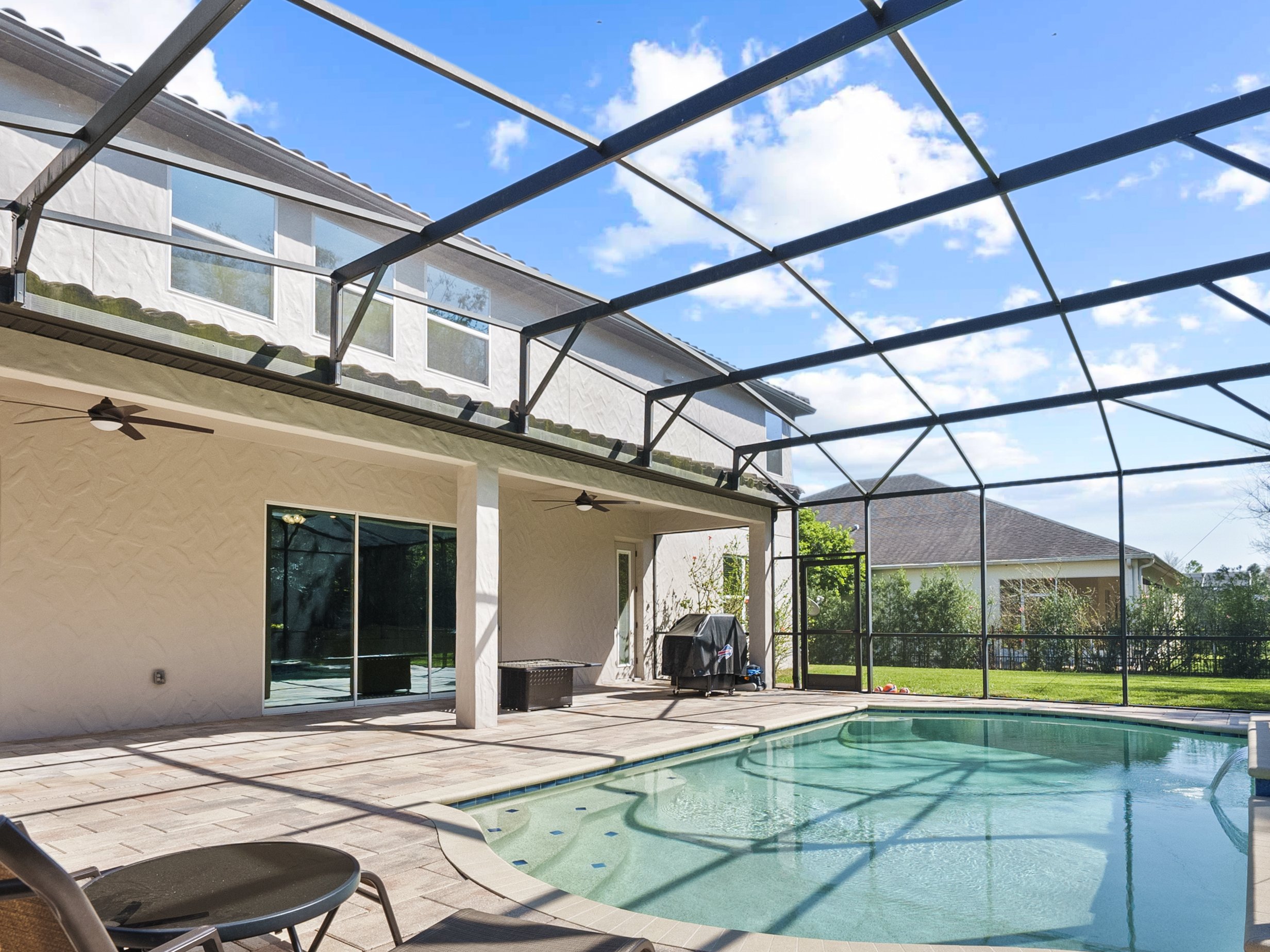 Screened-in backyard pool area with a patio, featuring ceiling fans and lounge chairs, adjacent to a two-story house. The area is enclosed by a screened frame, and the pool features steps leading into it. The house has sliding glass doors and windows
