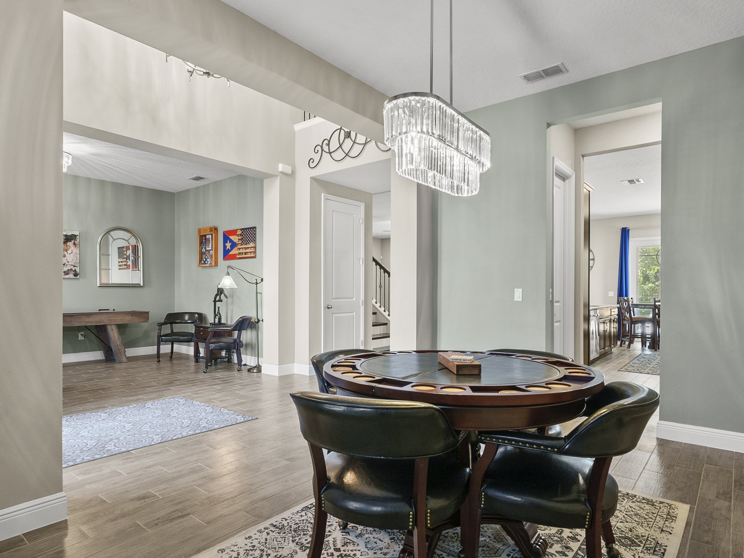 Interior of a residential home featuring a modern game room. In the foreground, a round poker table with chairs is set on a patterned rug. A hanging chandelier provides lighting. In the background, a hallway leads to a dining area with blue curtains,
