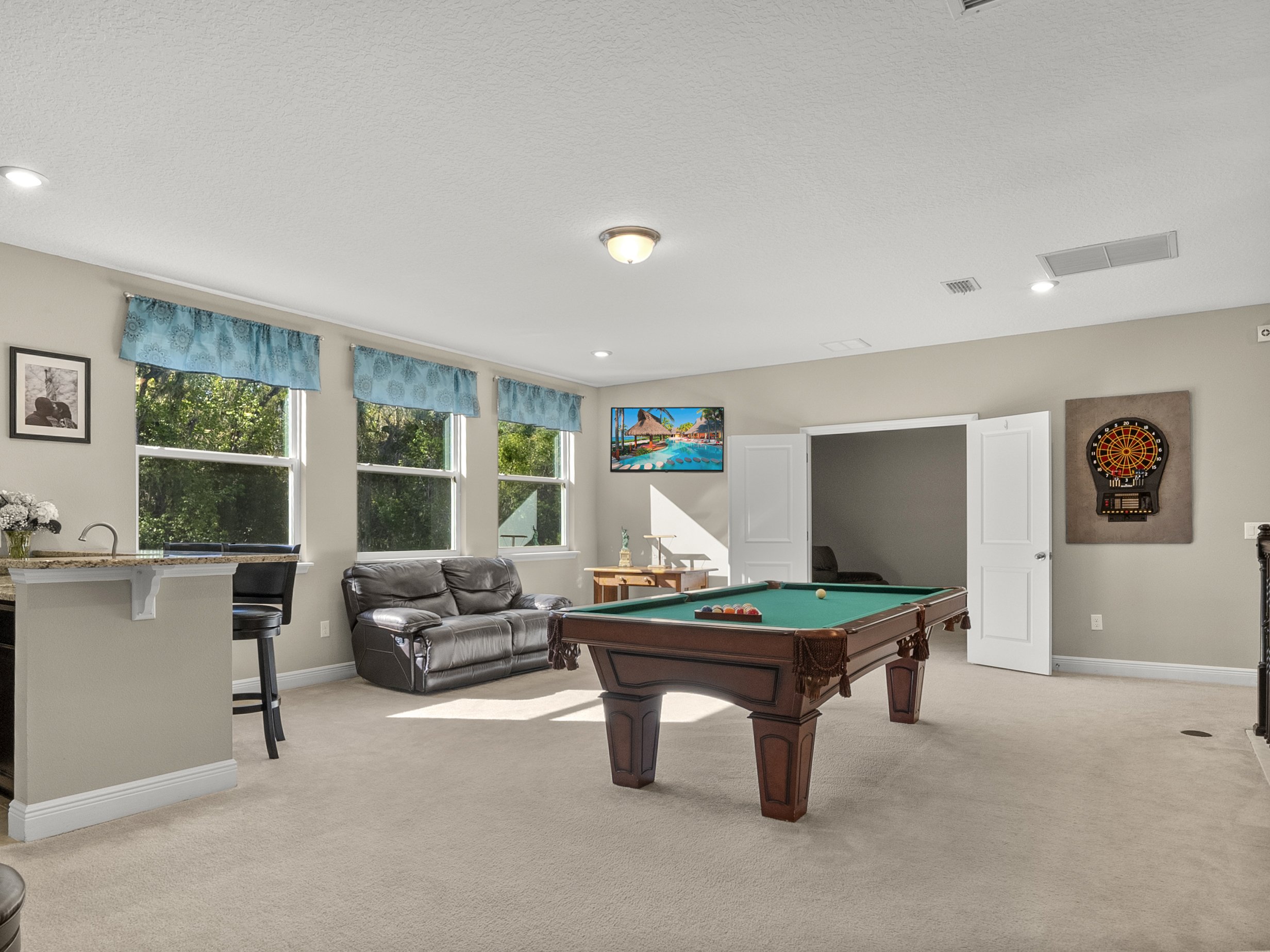 Interior of a game room with a pool table, dartboard, and seating area. The room features large windows with blue curtains, a leather sofa, a bar counter with stools, and decorative wall art. The floor is carpeted and the space is brightly lit by nat