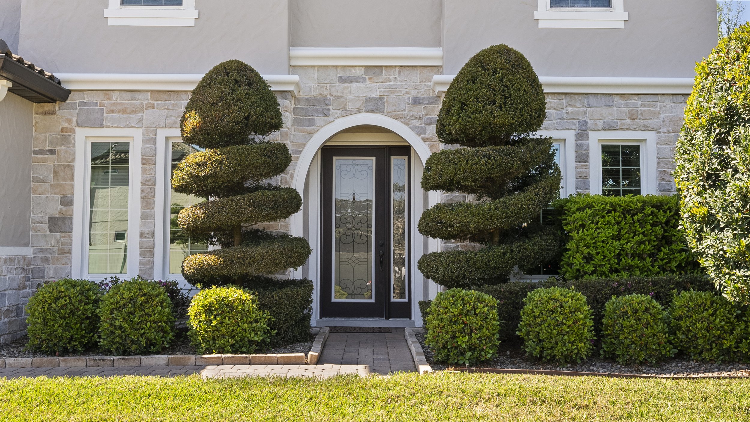 Front view of a house with a stone facade, featuring a decorative glass door and symmetrical topiary shrubs on each side. Windows with white trim are visible, surrounded by neatly trimmed bushes.