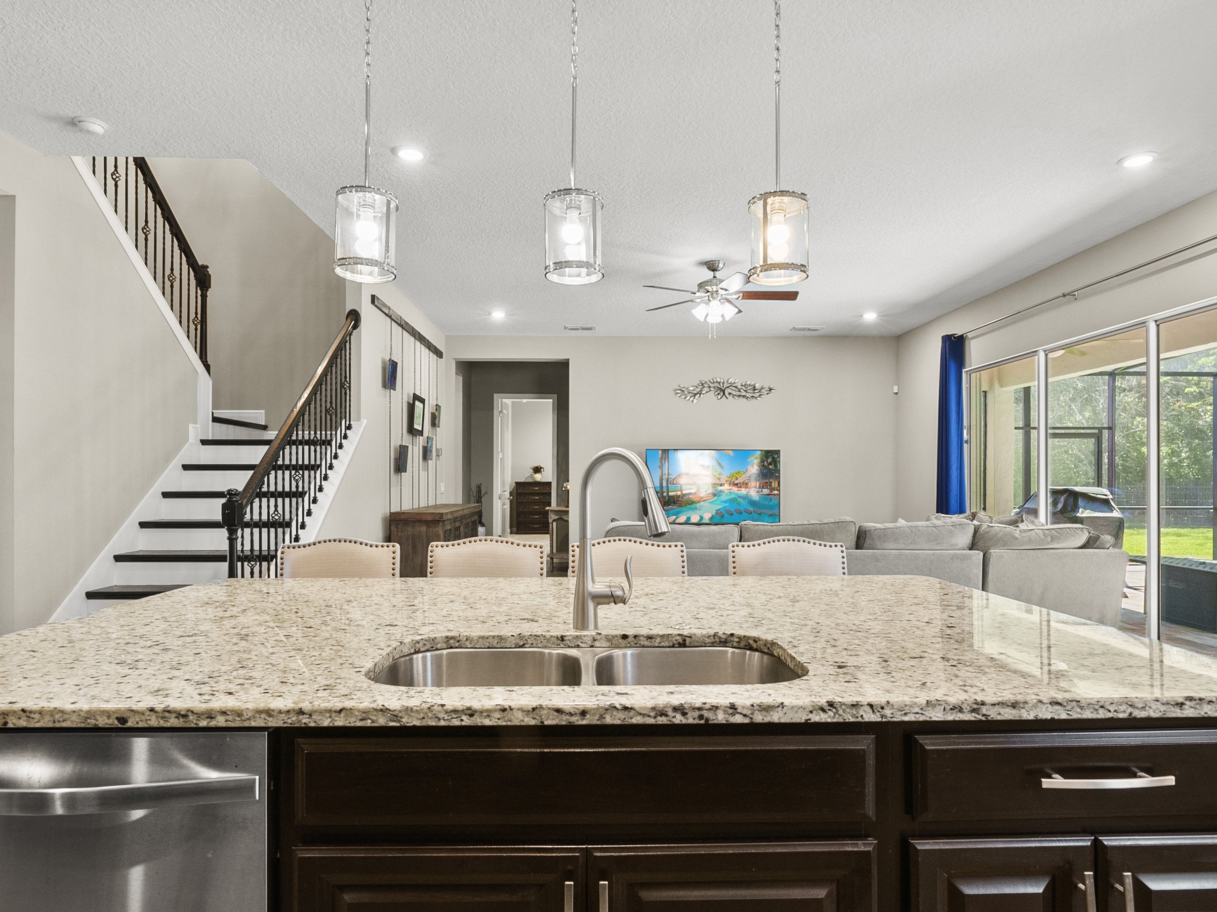 Modern open-concept living space with a granite kitchen island, pendant lighting, sectional sofa, wall-mounted TV, and large sliding doors leading to a patio.