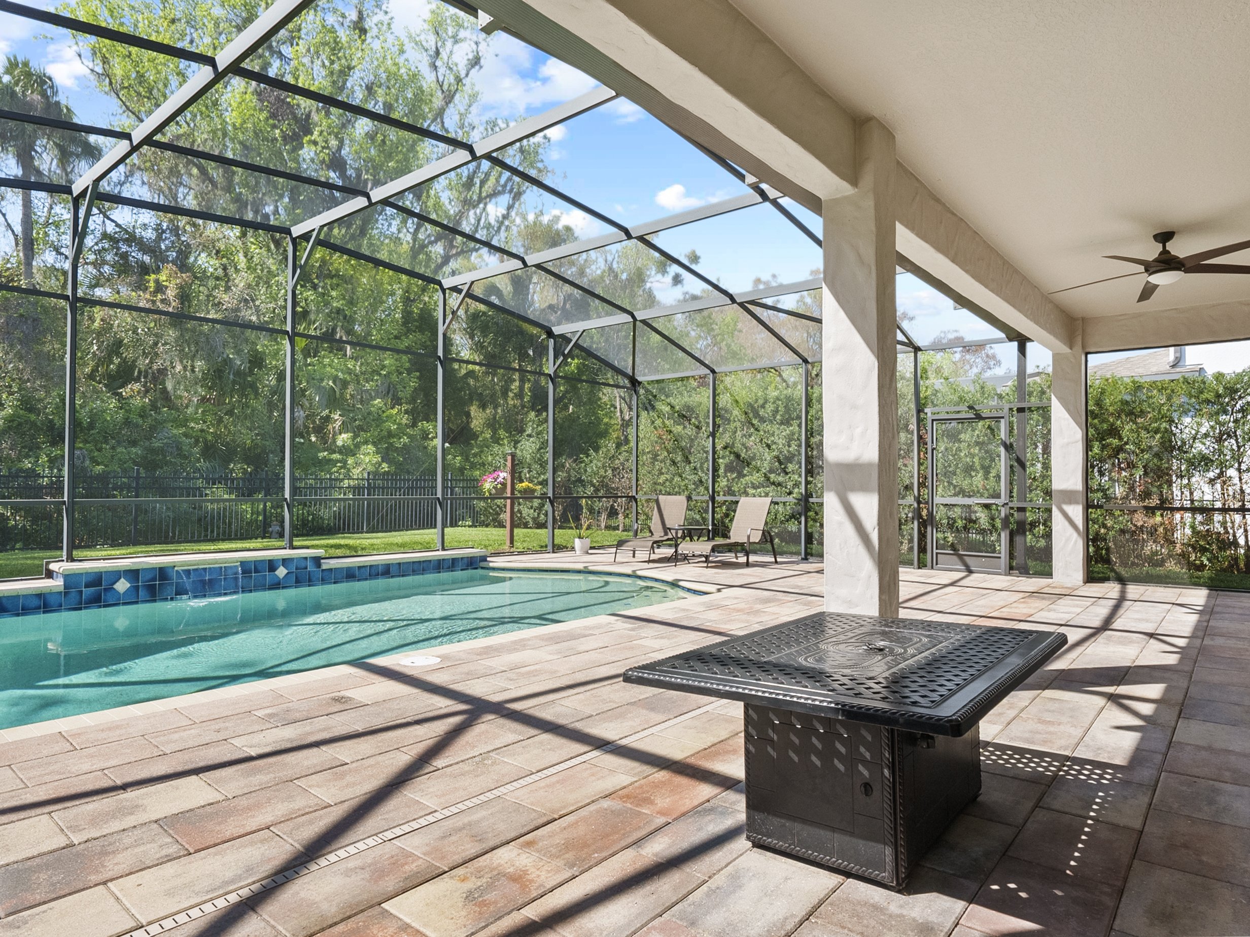 Screened-in outdoor patio with a pool and lounge chairs, surrounded by greenery and trees, featuring a ceiling fan and a metal fire pit table.