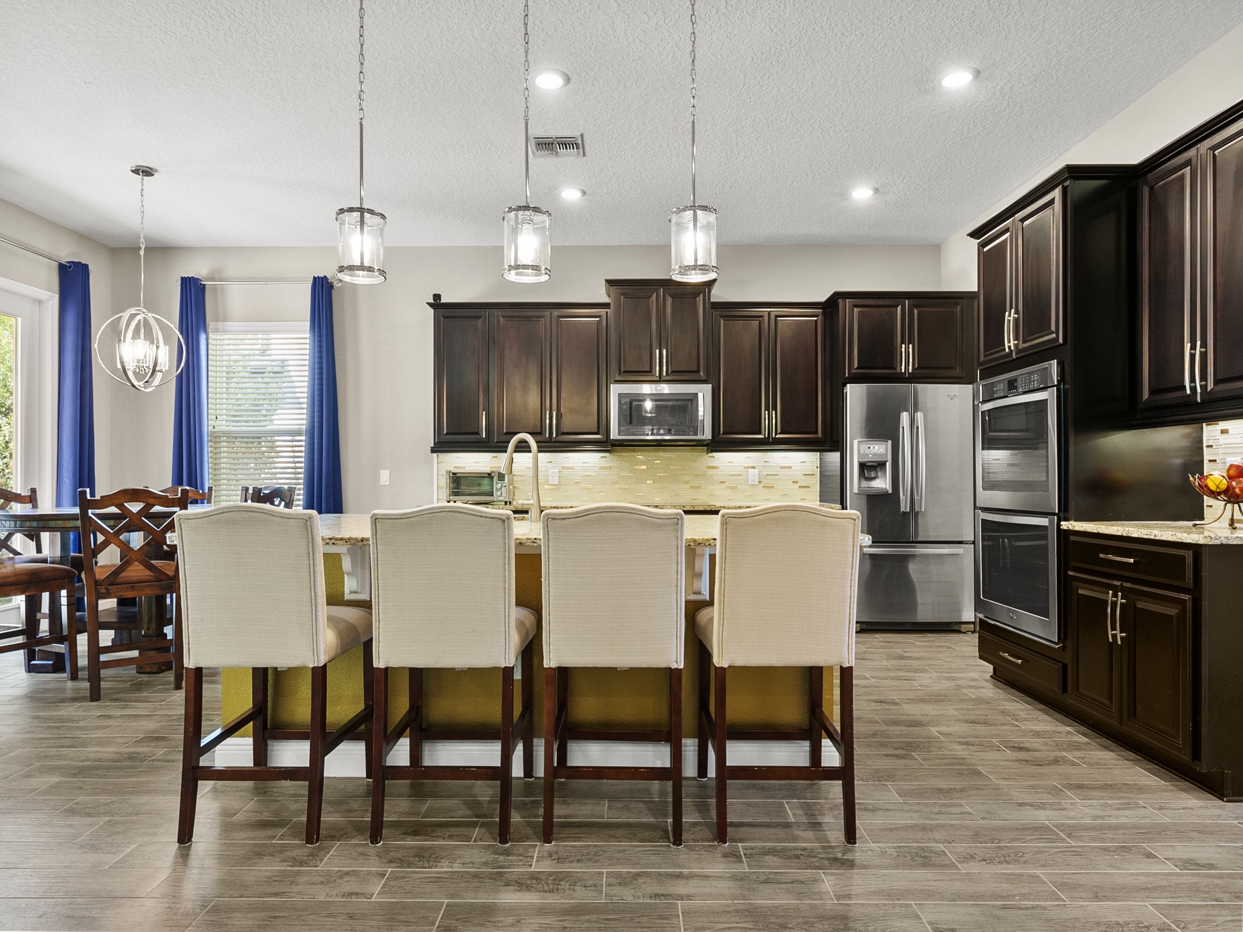 Modern kitchen with a central island, beige bar stools, dark wooden cabinets, stainless steel appliances, and pendant lighting. Dining area with a round table and blue curtains is visible in the background.