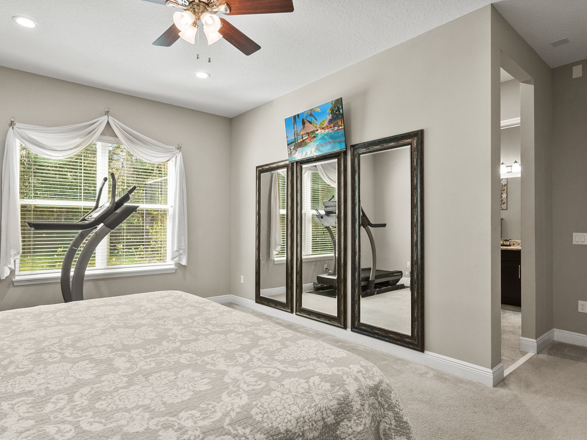 Bedroom with treadmill, ceiling fan, large windows with light curtains, bed with patterned comforter, and large framed mirrors on one wall.