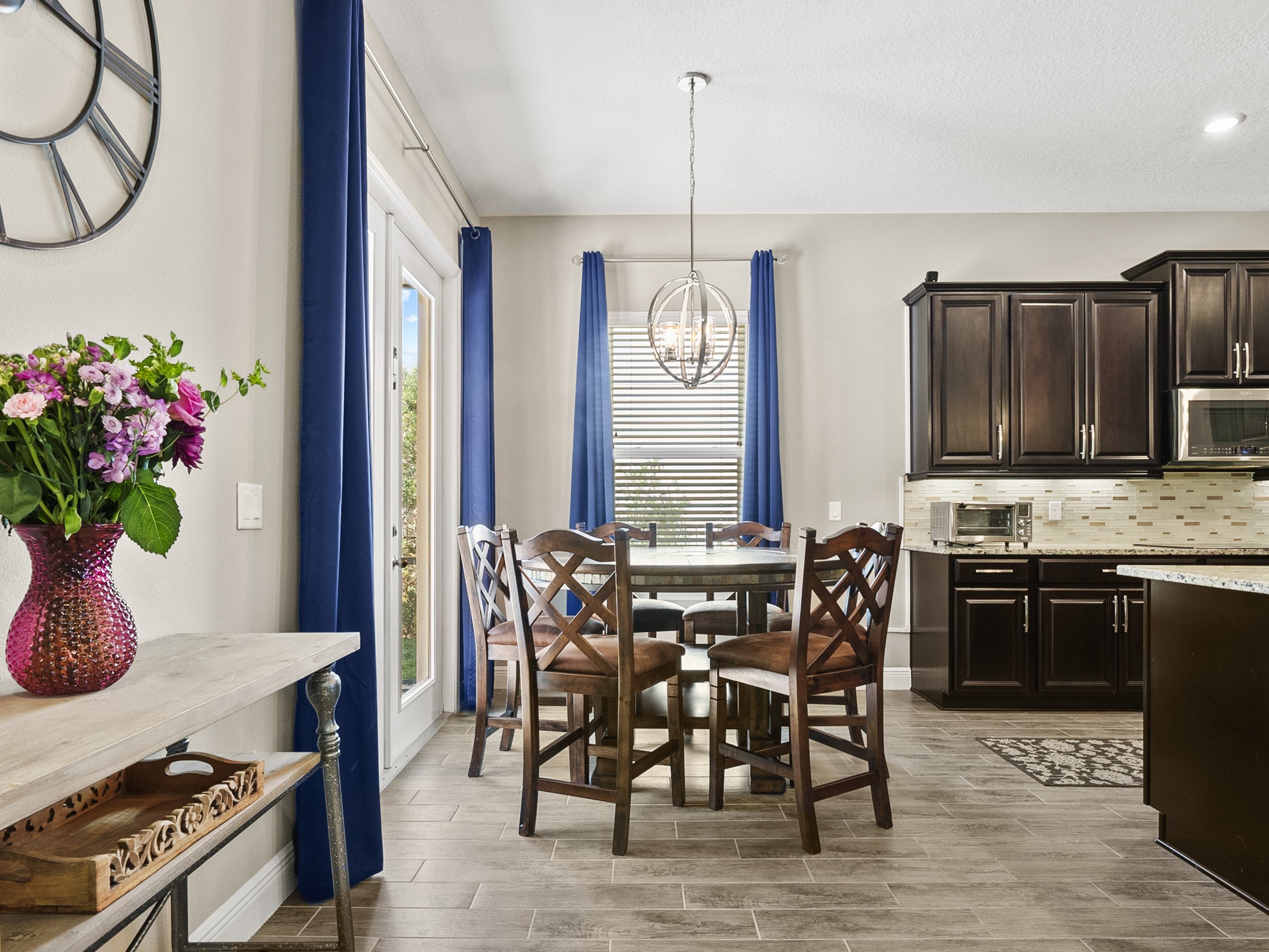 Modern kitchen and dining area with dark wooden cabinets, granite countertops, and a round dining table with chairs. A large clock is on the wall, and a vase with pink and purple flowers is on a nearby table. Blue curtains frame the windows.