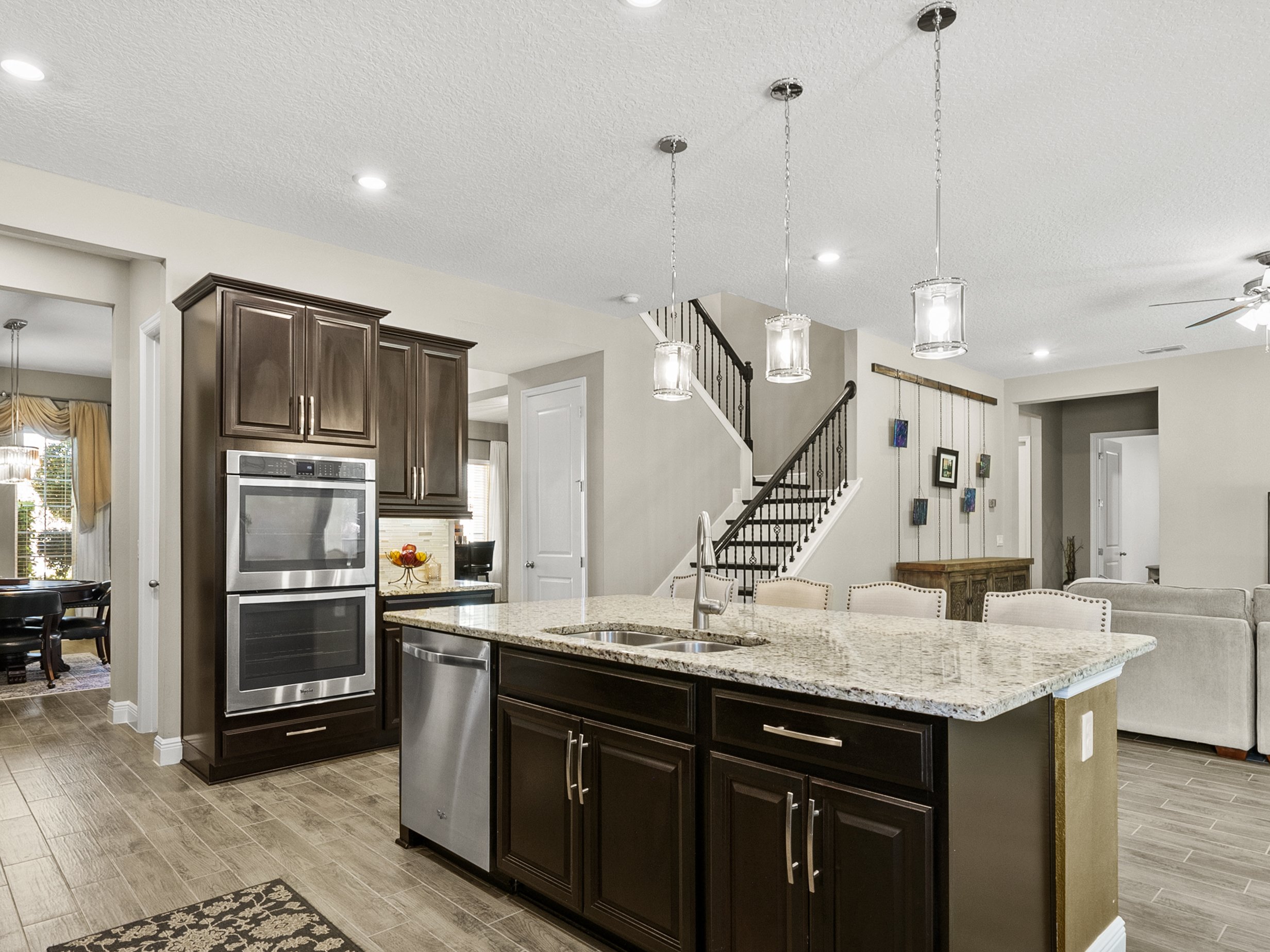 Modern kitchen interior with granite countertops, a central island, stainless steel appliances, dark wood cabinets, pendant lighting, and adjacent living areas with staircase visible.