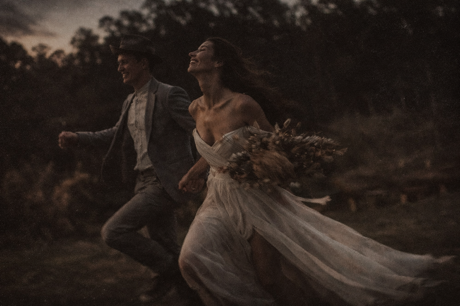 Bride and groom running hand in hand through a field at dusk, with a flowing dress and wild bouquet.