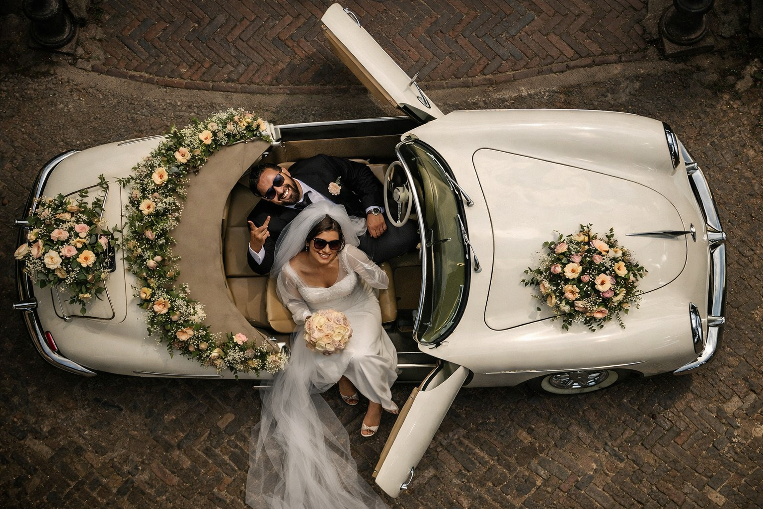 Photo de mariage vue du dessus d’un couple de mariés dans une voiture blanche vintage décorée de fleurs sur une cour en briques.