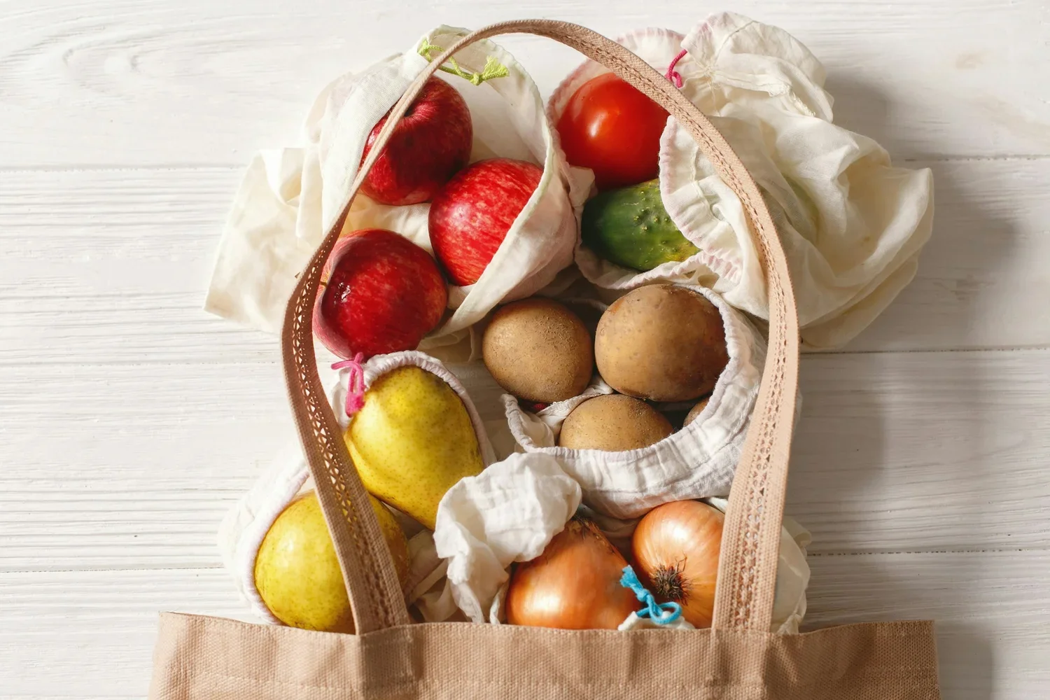 A reusable shopping bag with apples, potatoes, a cucumber, a yellow pear, and onions on a white wooden surface.