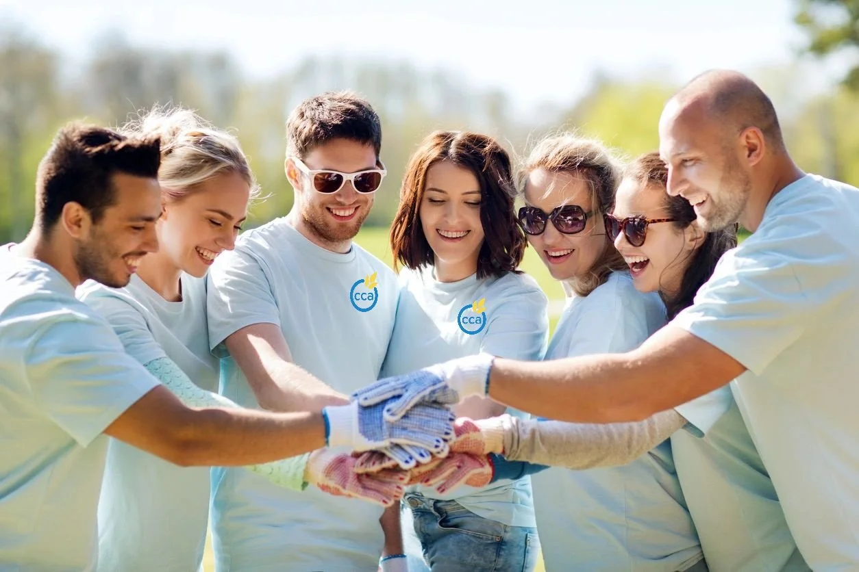 Group of people outdoors wearing white shirts with a blue and yellow logo, smiling, and putting their hands together in a team gesture.