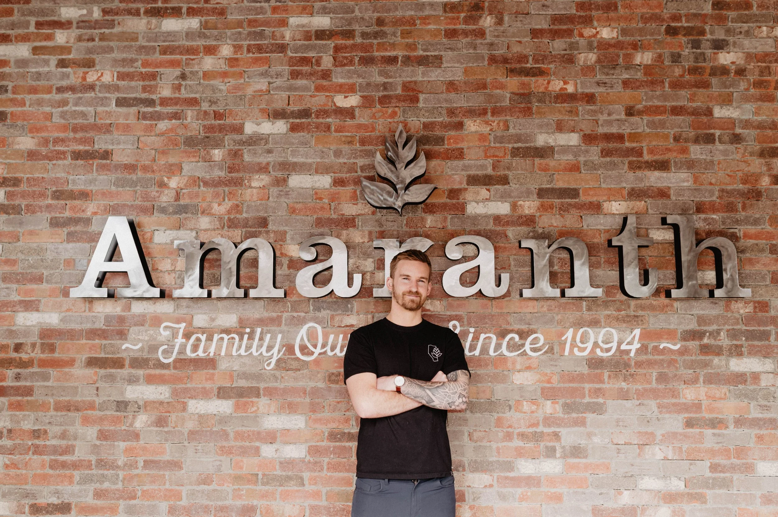 A man with a beard and tattoos standing with arms crossed in front of a brick wall with the word 'Amaranth' and the phrase 'Family Owned Since 1994' on it.