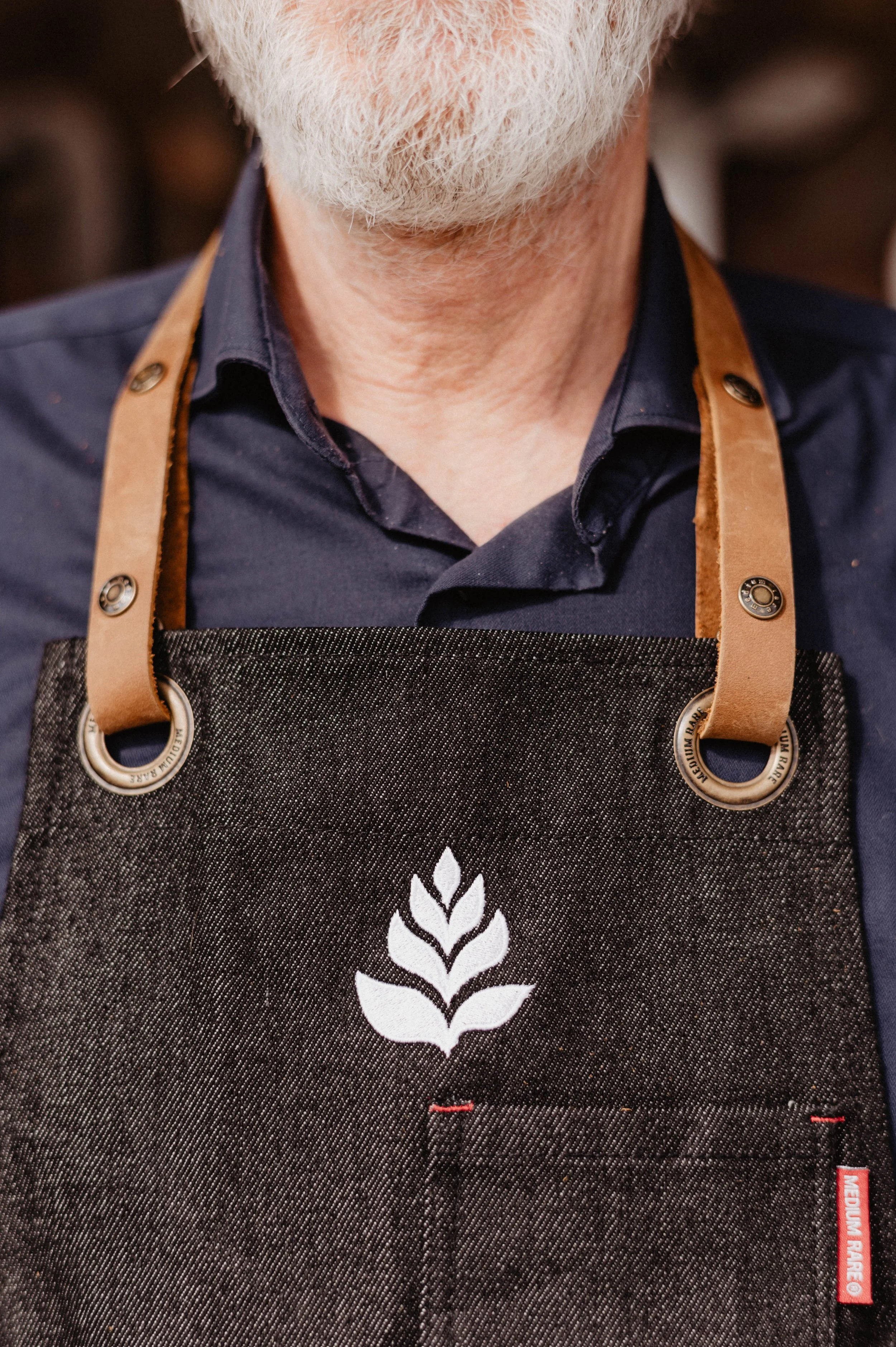 Close-up of a man with a white beard wearing a navy blue shirt and a black apron with a white logo, brown leather strap, and metal rings.