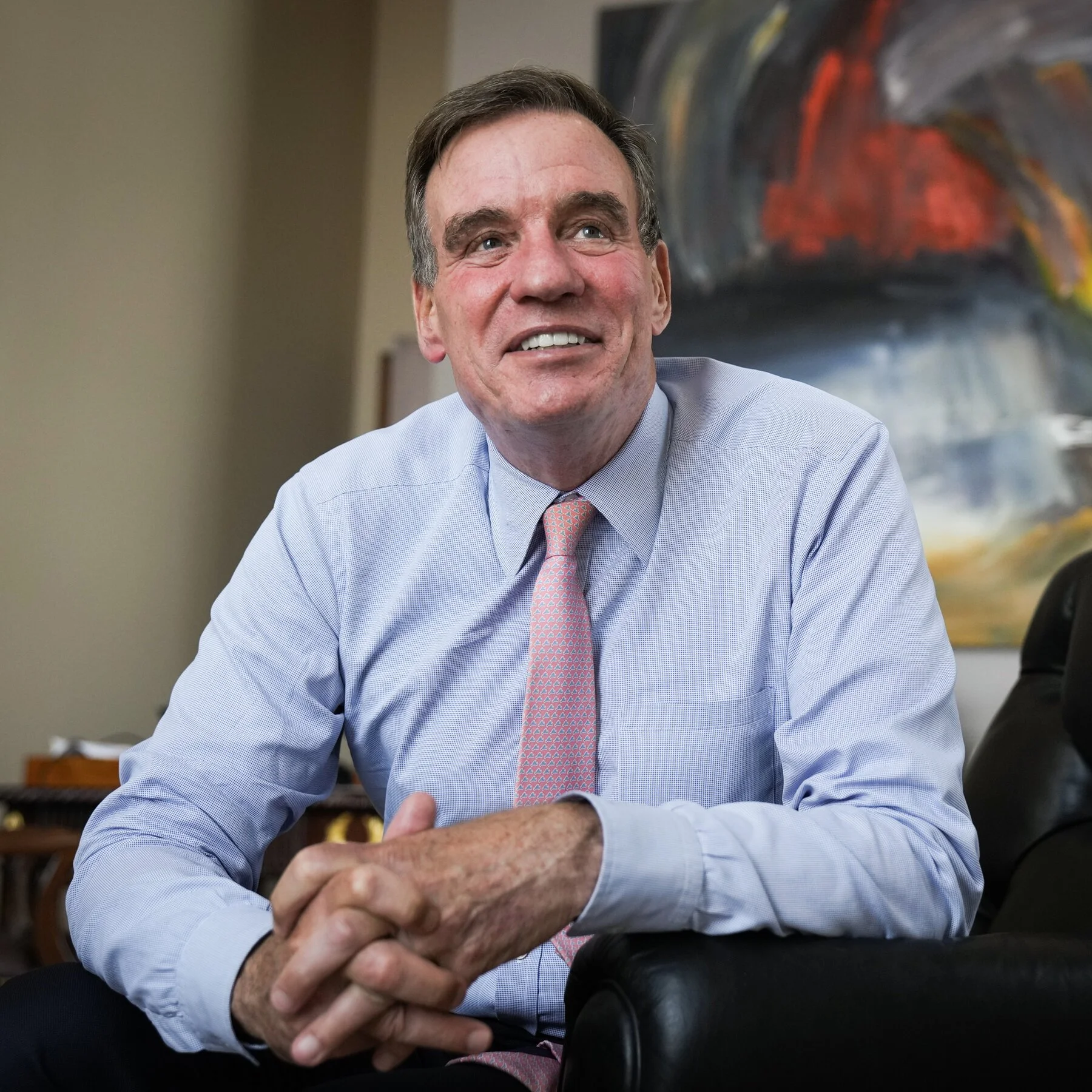 A man in a light blue dress shirt and pink patterned tie, sitting in an office, smiling and clasping his hands. There is an abstract painting in the background.