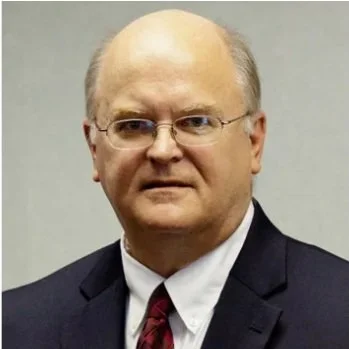 A middle-aged man in a dark suit, white shirt, and patterned tie, with glasses and light hair, posing for a portrait against a plain background.