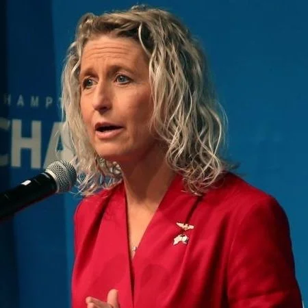 A woman with curly gray hair wearing a red blazer and a pin, speaking into a microphone against a blue background.