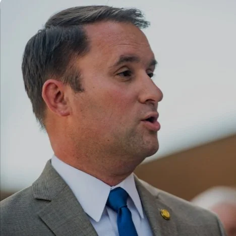 A man in a gray suit with a blue tie speaking, profile view, outdoor setting.