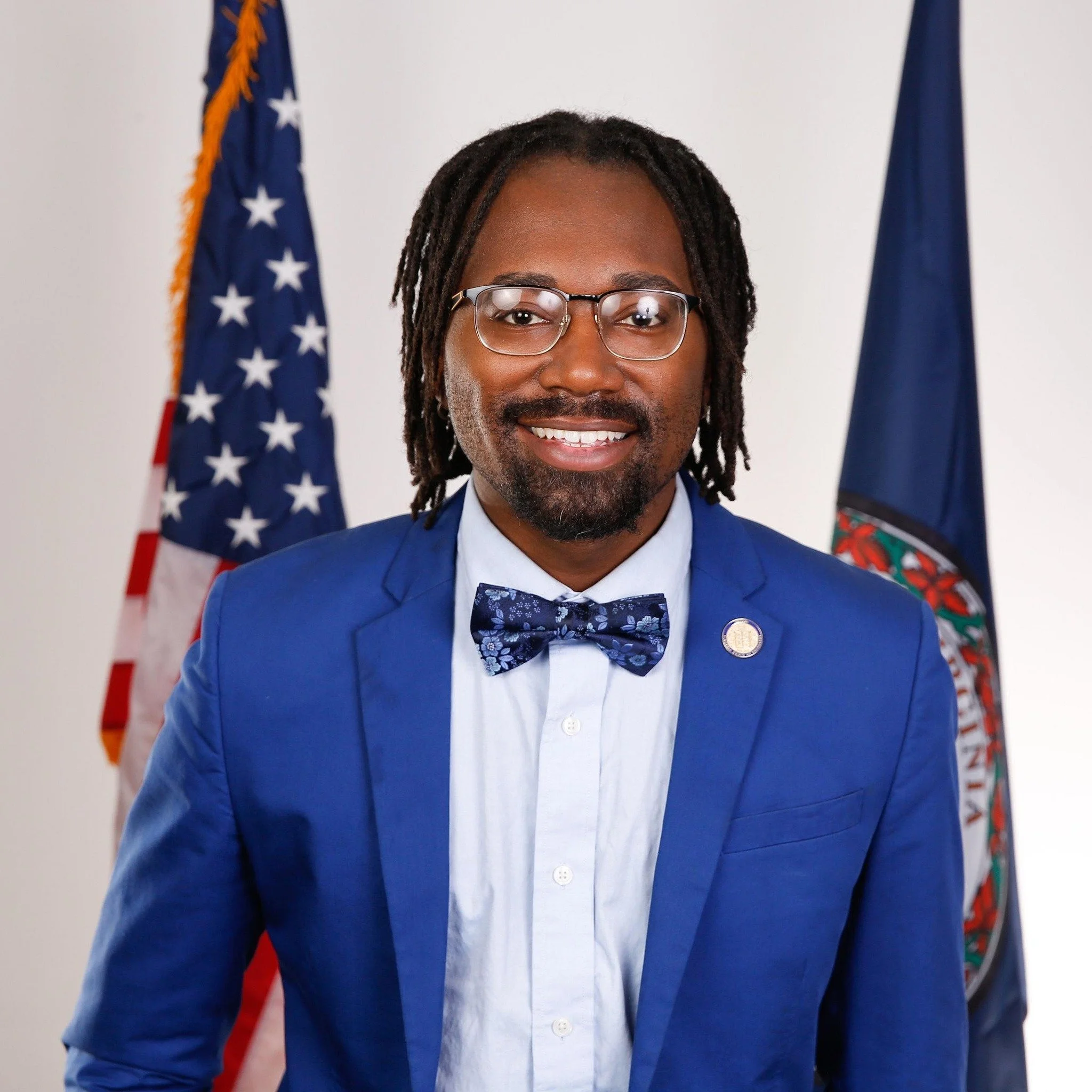 A man with glasses and dreadlocks wearing a blue suit, dress shirt, and bowtie, standing in front of American and State flags, smiling.