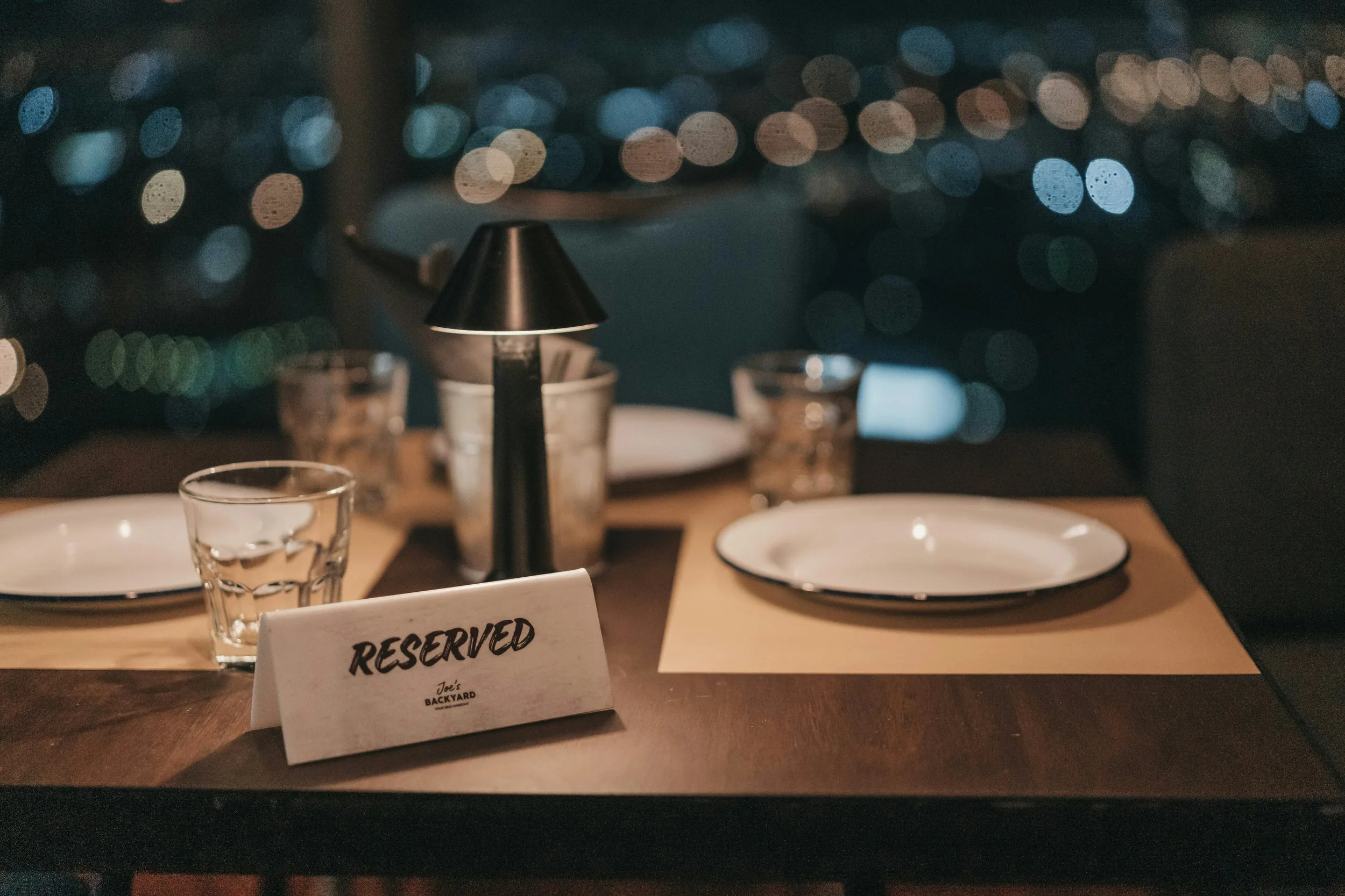 A restaurant table setting with a small lamp, empty plates, glasses, and a first in line 'RESERVED' sign, with a blurred background of bokeh lights.