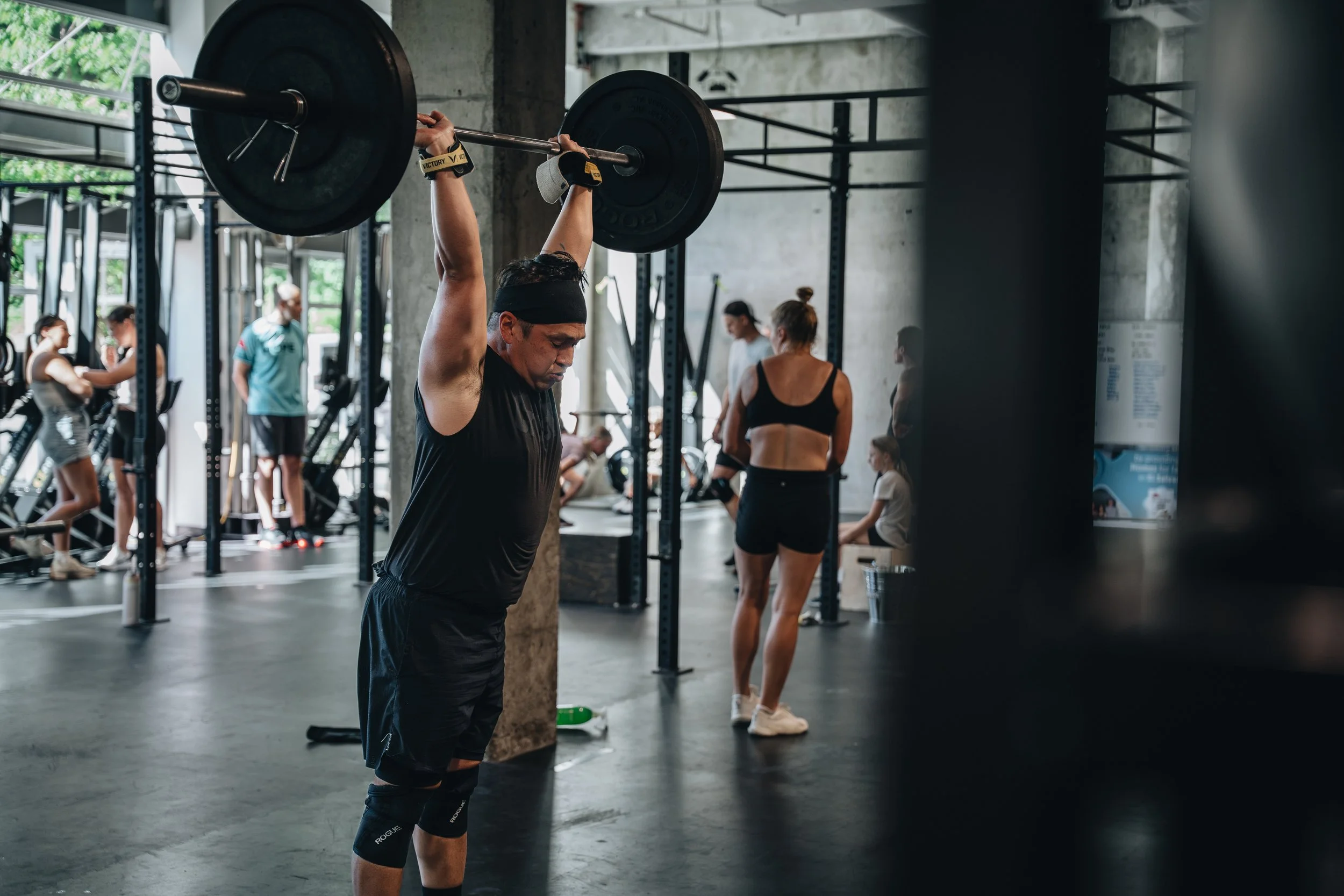 A man lifting a barbell overhead in a gym, with other people working out in the background.