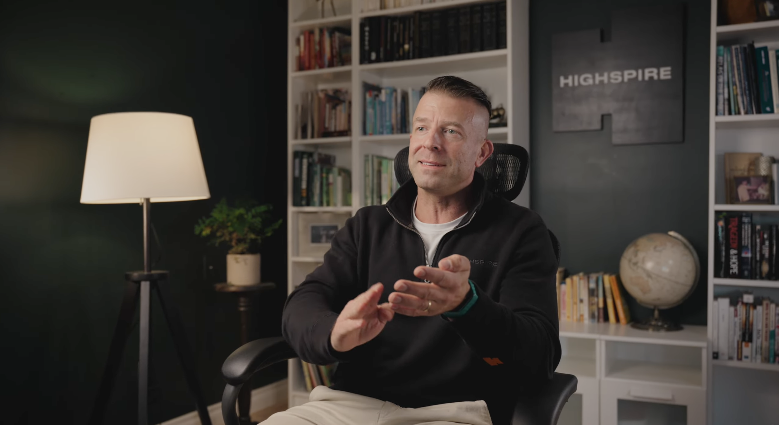 A man sitting in a black office chair in a room with a dark green wall, a bookshelf filled with books, a globe, and a table lamp. The man is wearing a black jacket and is gesturing with his hands.