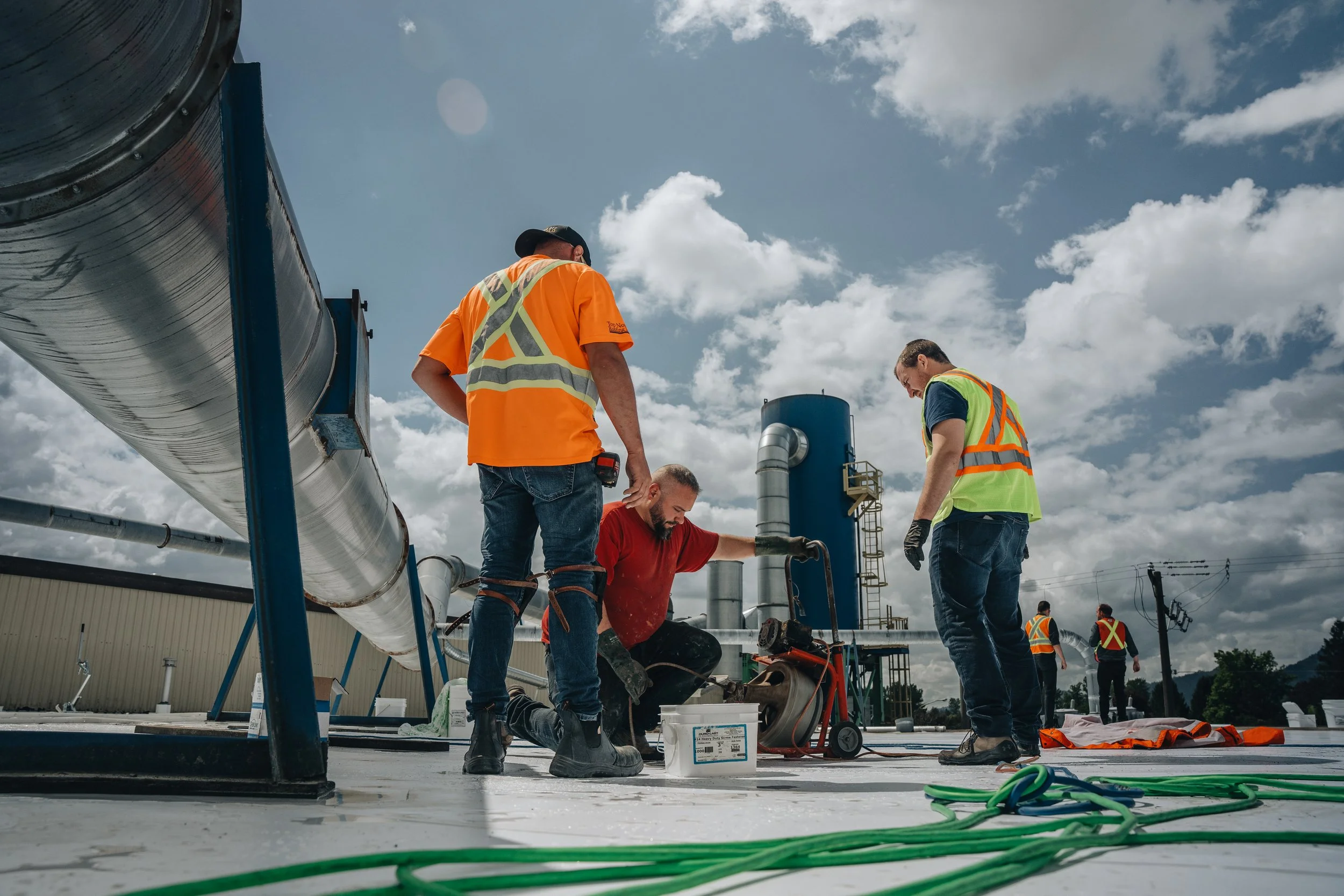 Construction workers working on a rooftop with large industrial equipment and pipes under a partly cloudy sky.