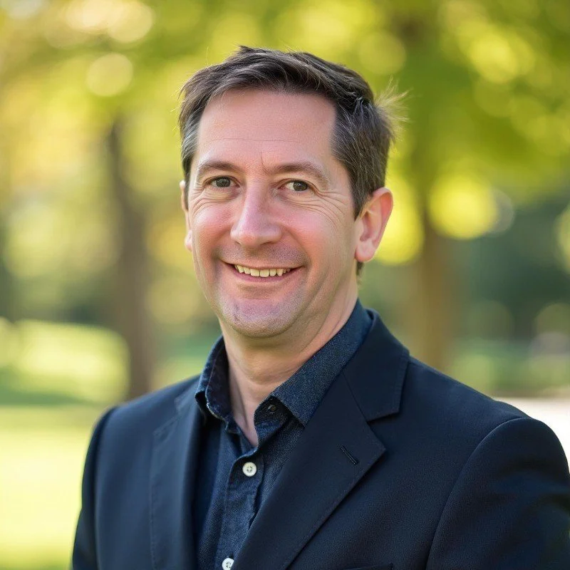 A man with short brown hair in a black blazer and dark shirt outdoors on a sunny day, smiling, with blurred green trees in the background.