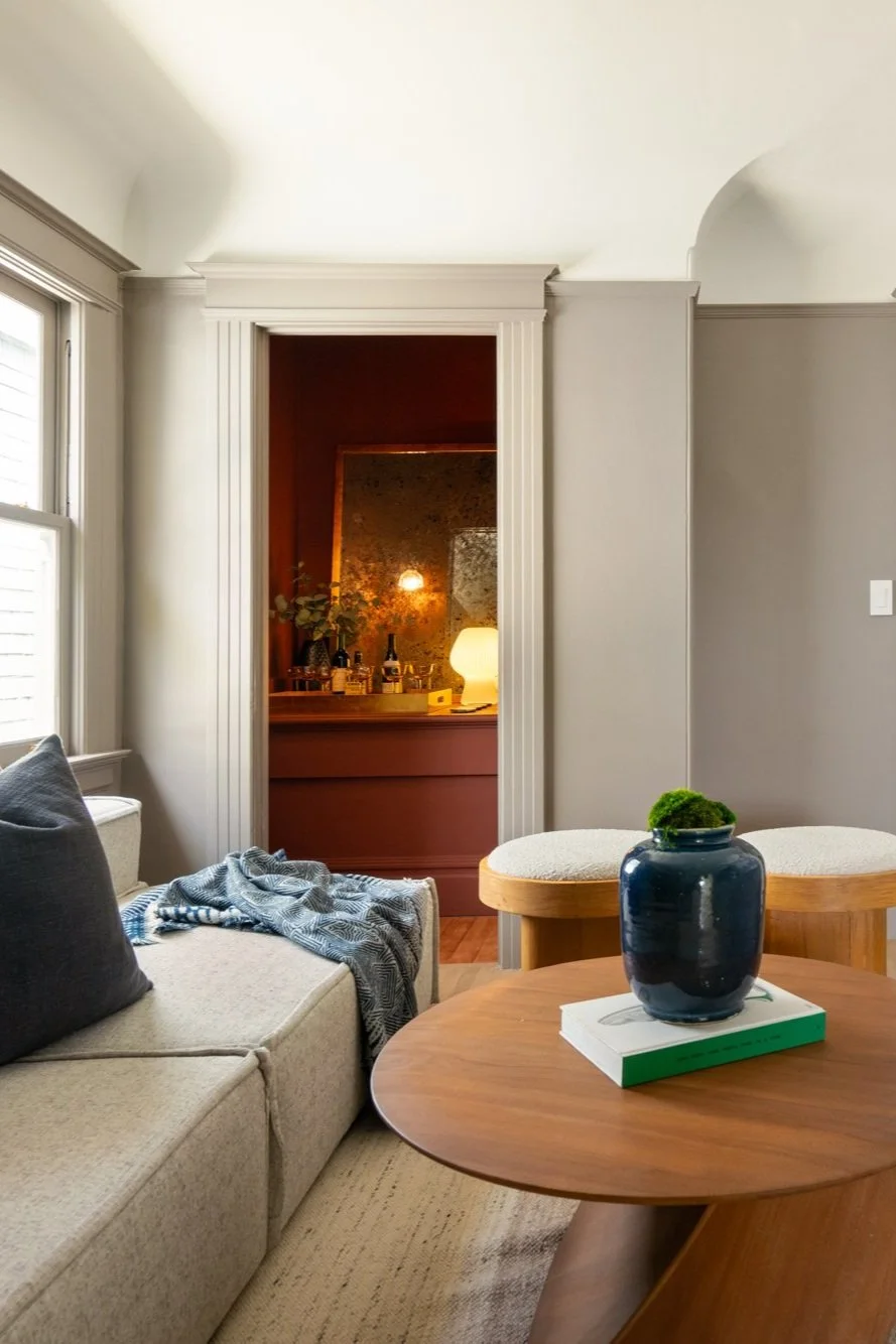 Cozy living room with a light gray sofa, blue pillow, and patterned throw blanket. Wooden coffee table with a blue vase and book. Two cushioned stools are nearby. The room opens into a warm brown space with a lit lamp and bottles on display.