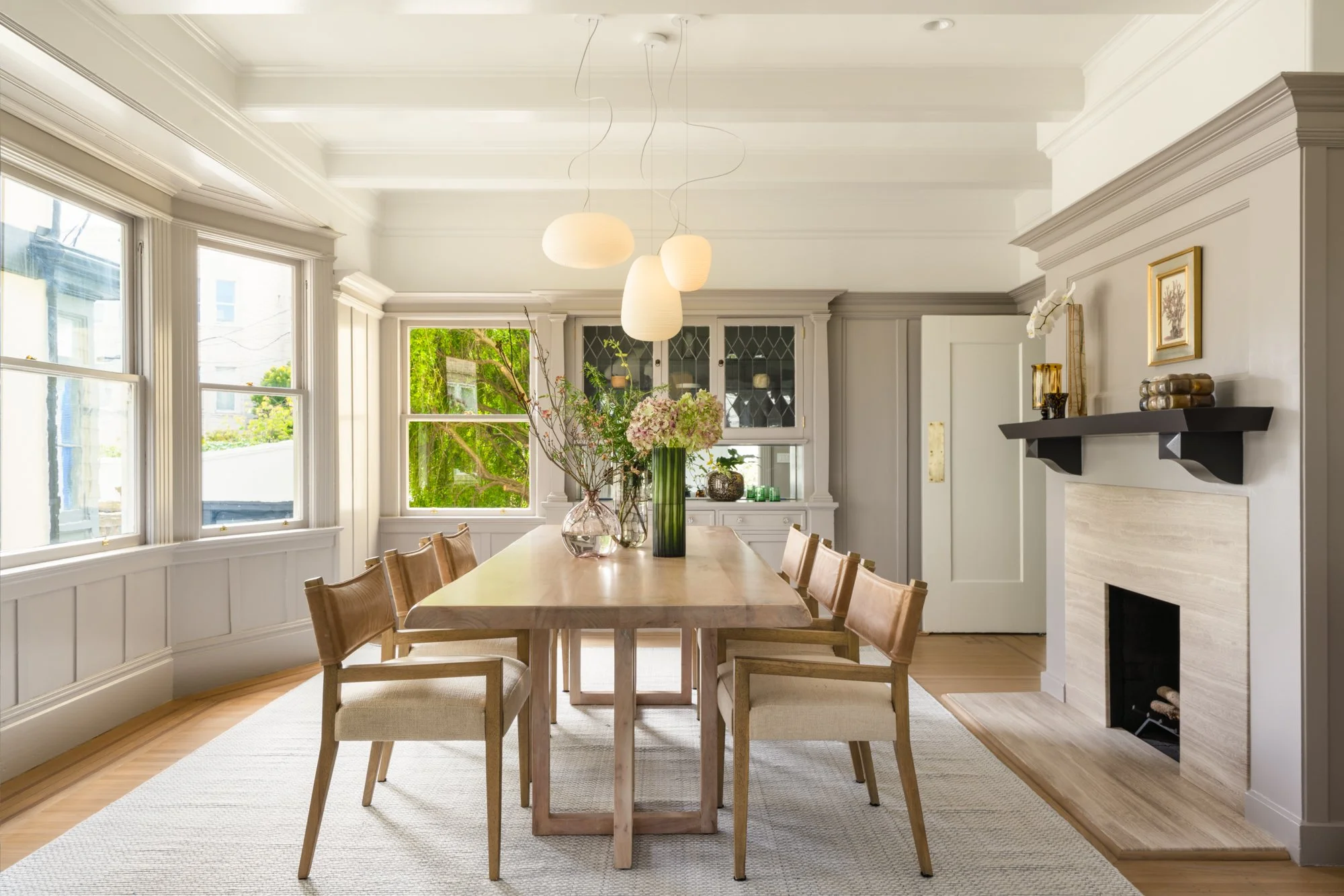 Elegant dining room featuring a wooden table with chairs, modern pendant lights, a fireplace, and a cabinet, with large windows providing natural light.