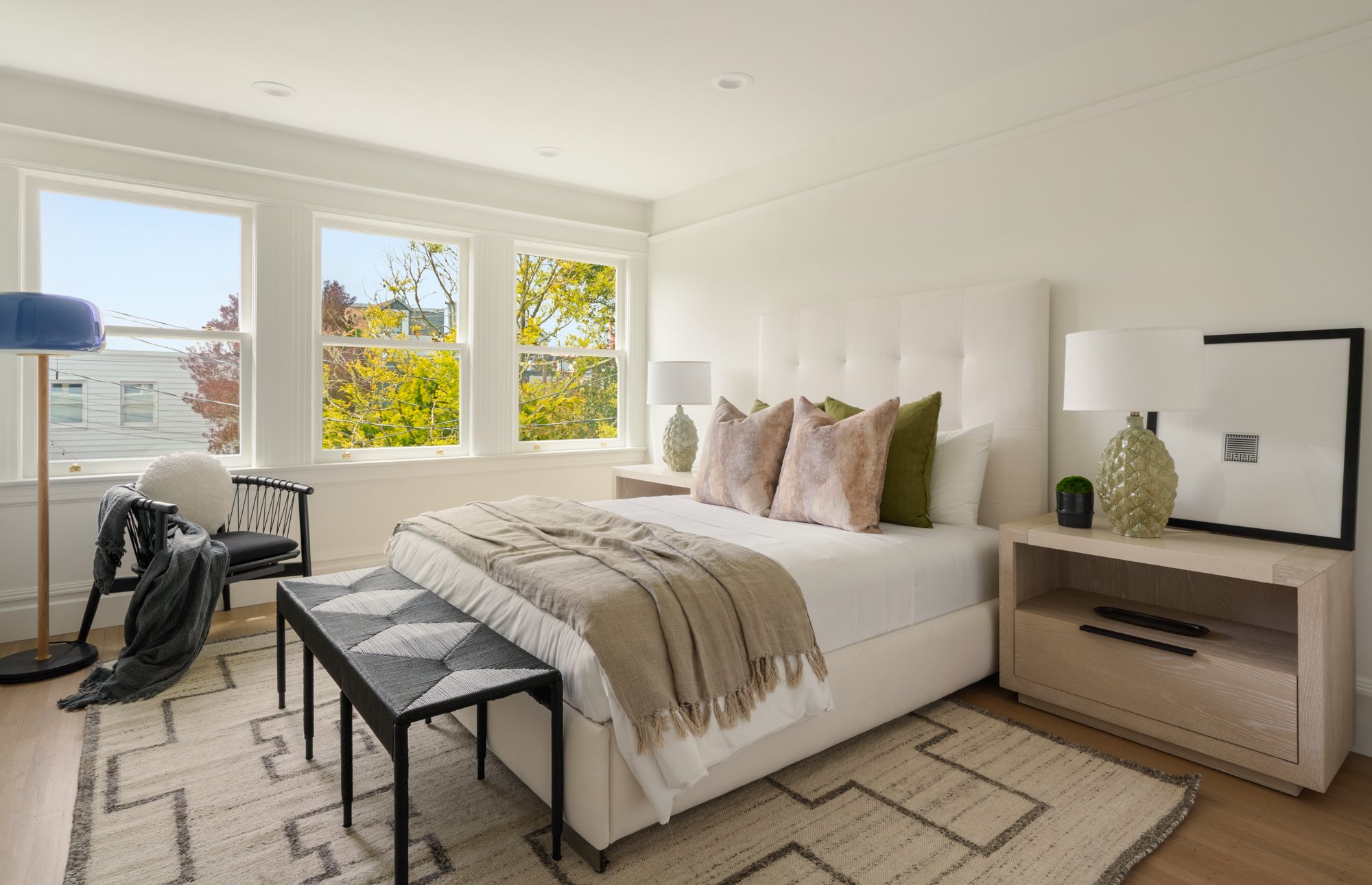 Modern bedroom with large windows, neutral decor, and a white bed with pillows. Features a light wood nightstand, table lamp, black chair with throw, and a decorative rug.