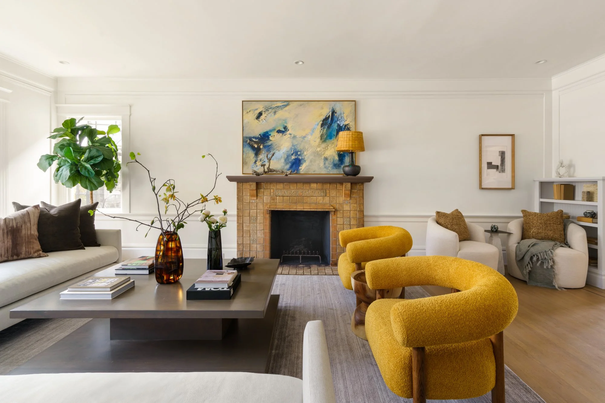 Modern living room with yellow chairs, a beige sofa, coffee table with vases, a brick fireplace, abstract artwork, and potted plant.