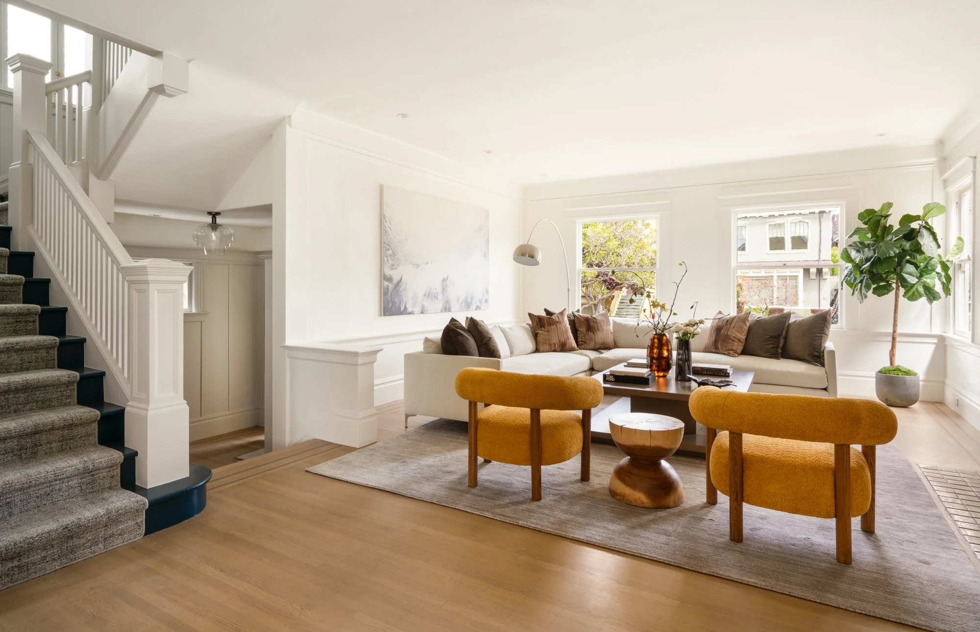 Modern living room with staircase, sectional sofa, yellow armchairs, wooden coffee table, large plant, and artwork.