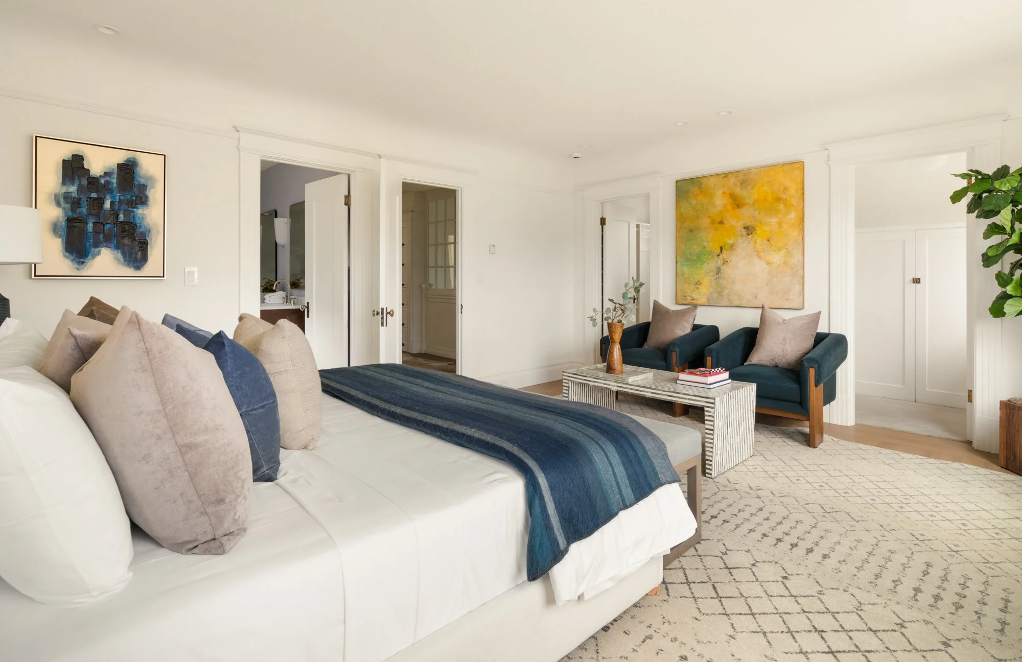 Modern bedroom with bed, pillows, and blue throw blanket, abstract art, two armchairs, coffee table, decorative vase, and indoor plant.