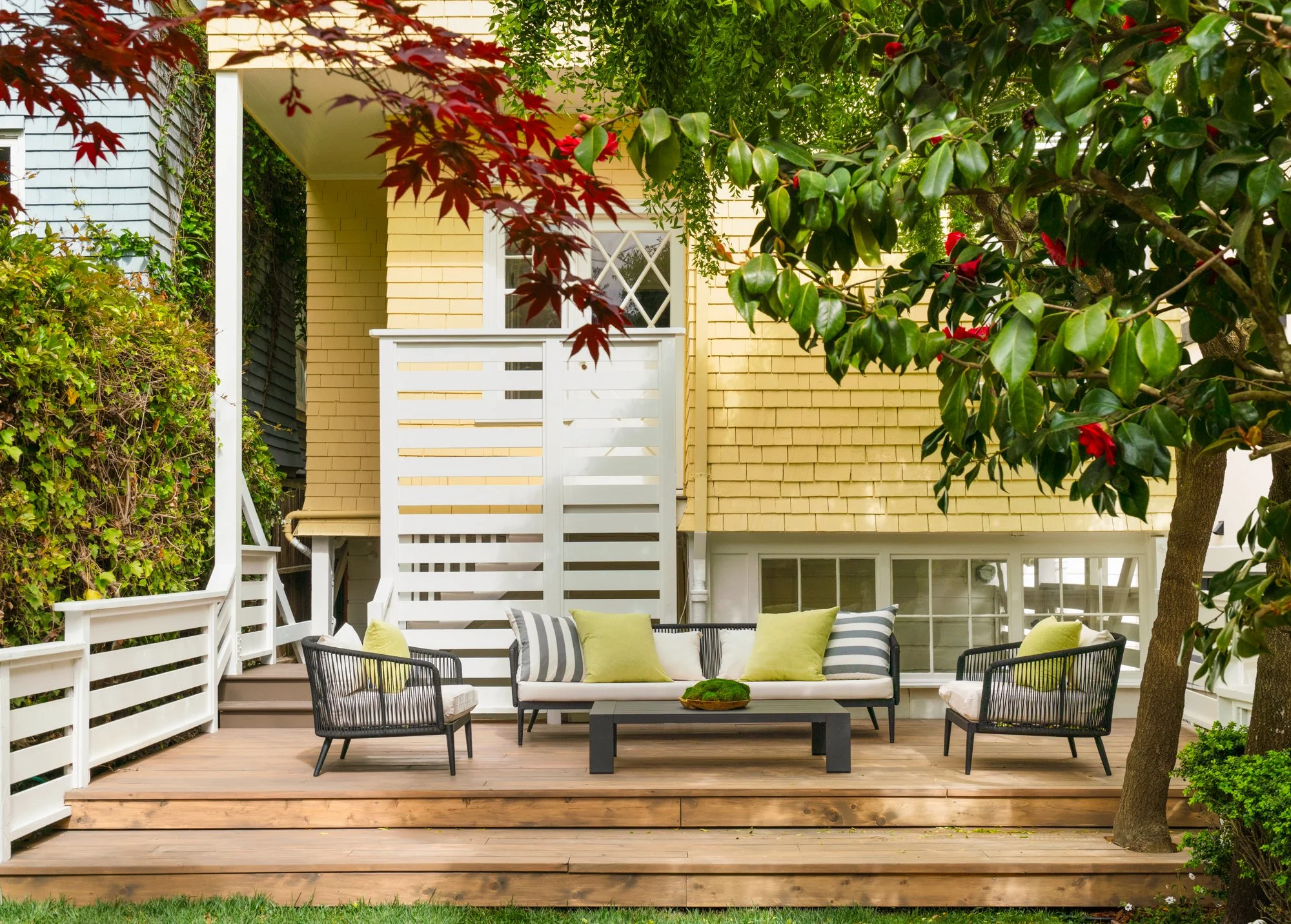 Outdoor patio with seating area featuring modern chairs, a coffee table, and striped cushions, surrounded by trees and a yellow house exterior.