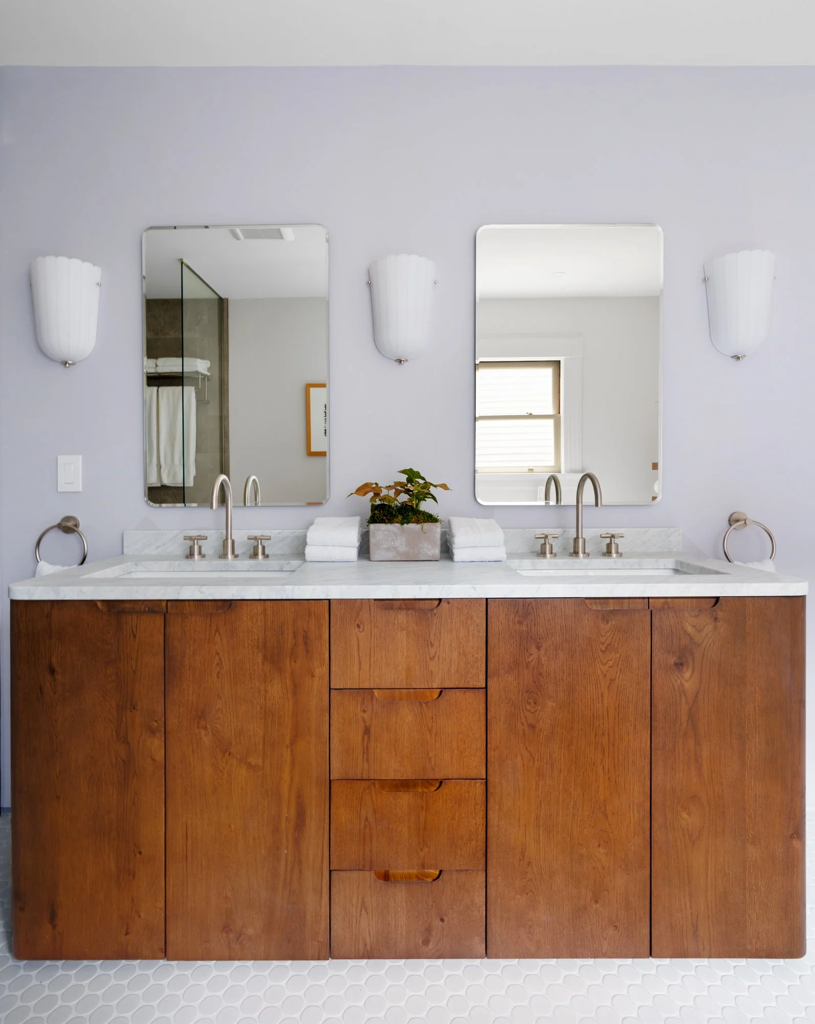 Bathroom vanity with double sinks, wooden cabinets, two mirrors, two wall sconces, stacked white towels, a potted plant, and a glass shower enclosure.