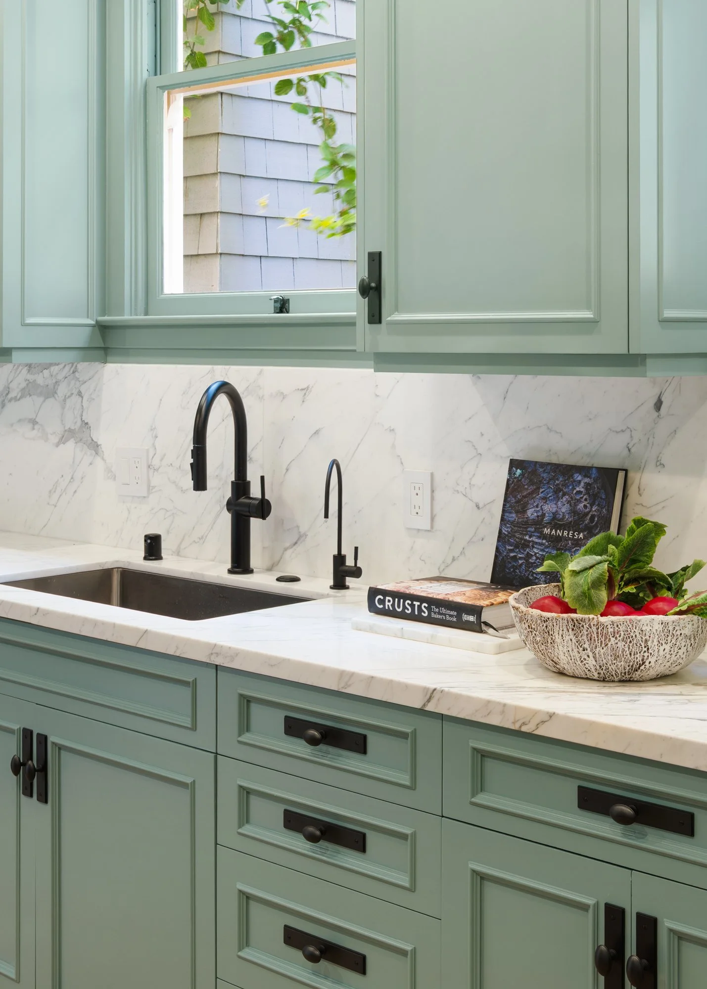 Kitchen with mint green cabinets, marble countertop, black faucets, a bowl of radishes, and cookbooks.