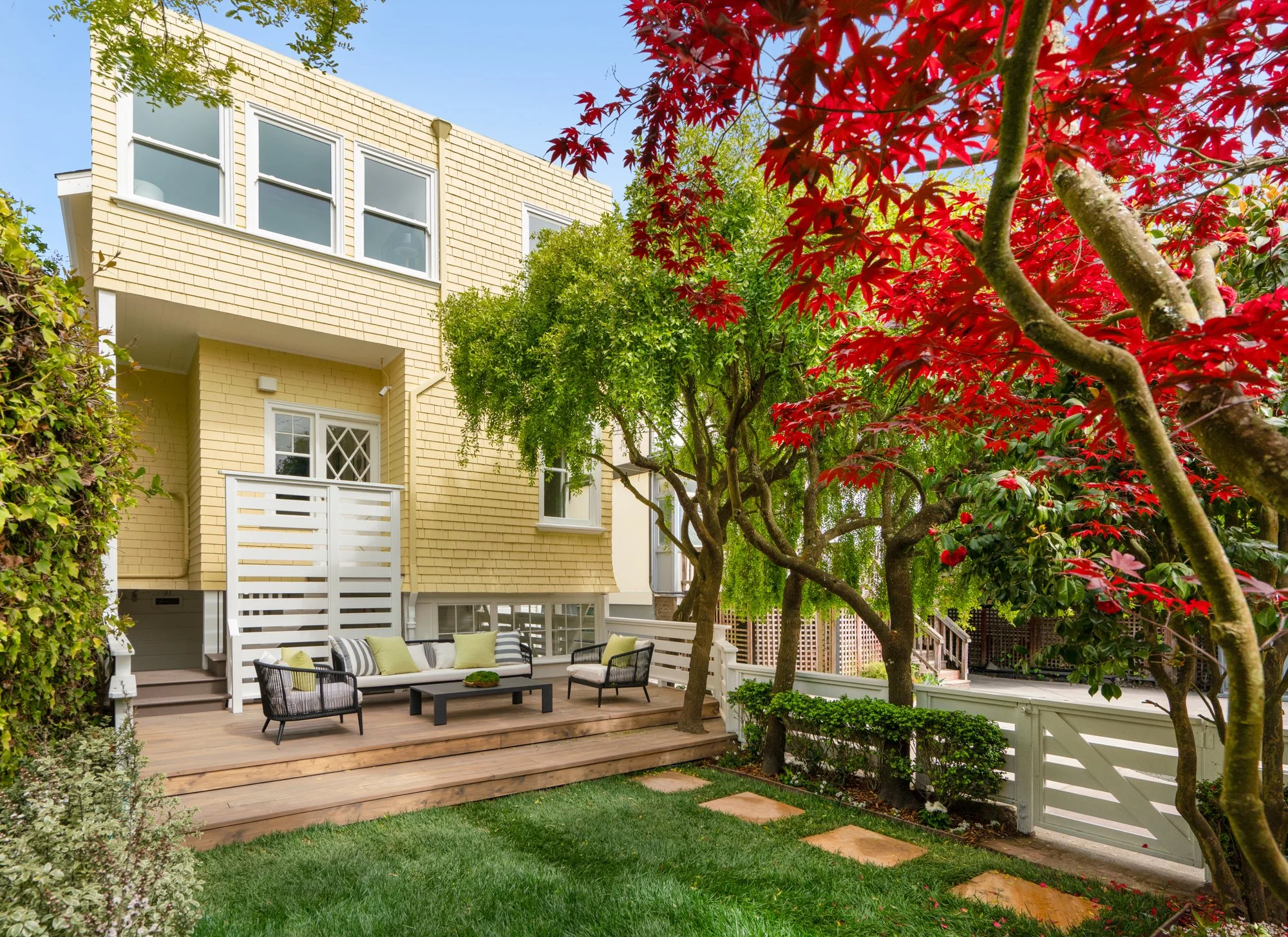 A backyard view of a two-story yellow house with a wooden patio. The patio features outdoor seating, including a couch and chairs with green and black cushions. The surrounding area has lush green and red trees, manicured grass, and stepping stones l