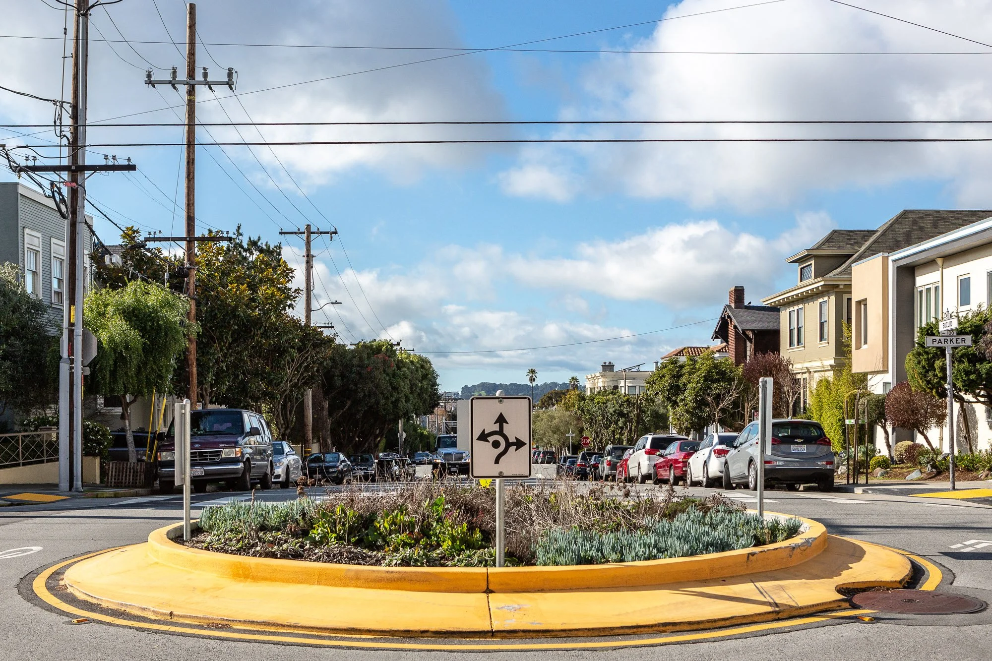 Street intersection with roundabout, traffic sign, parked cars, houses, and cloudy sky.