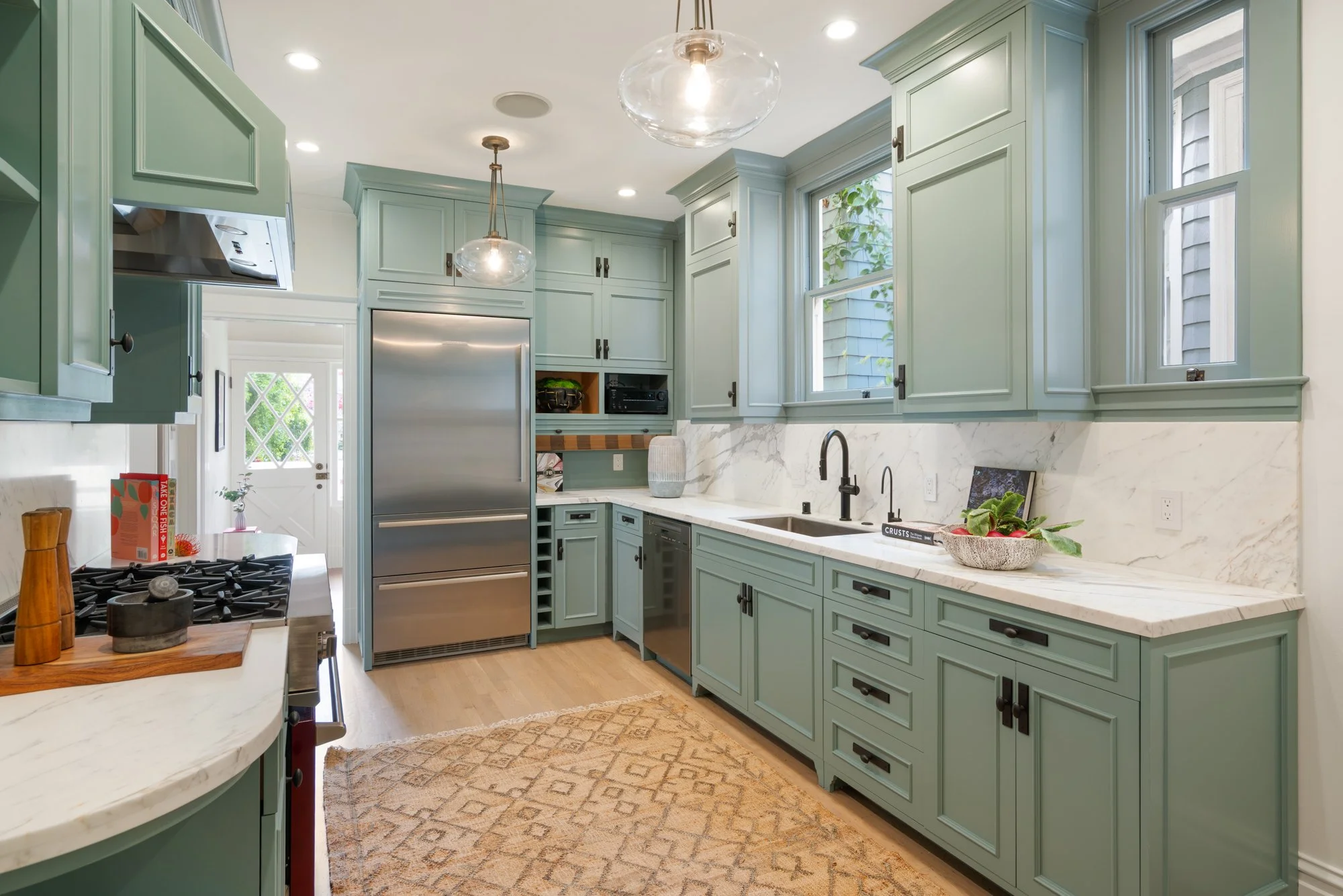 Modern kitchen with green cabinets, stainless steel appliances, marble countertops, a patterned rug, pendant lights, and a large window.
