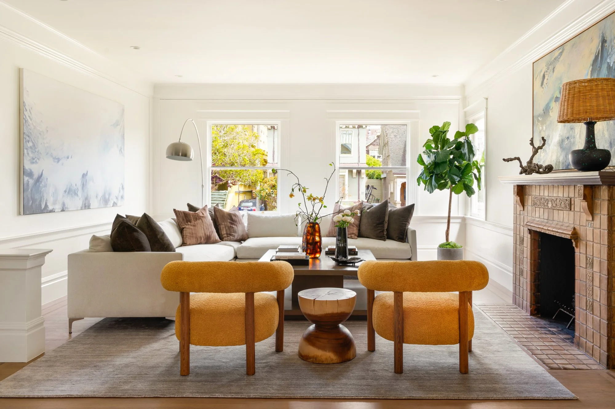 Stylish living room with beige sectional sofa, yellow armchairs, coffee table, fireplace, and large windows. Modern decor with indoor plant and abstract wall art.