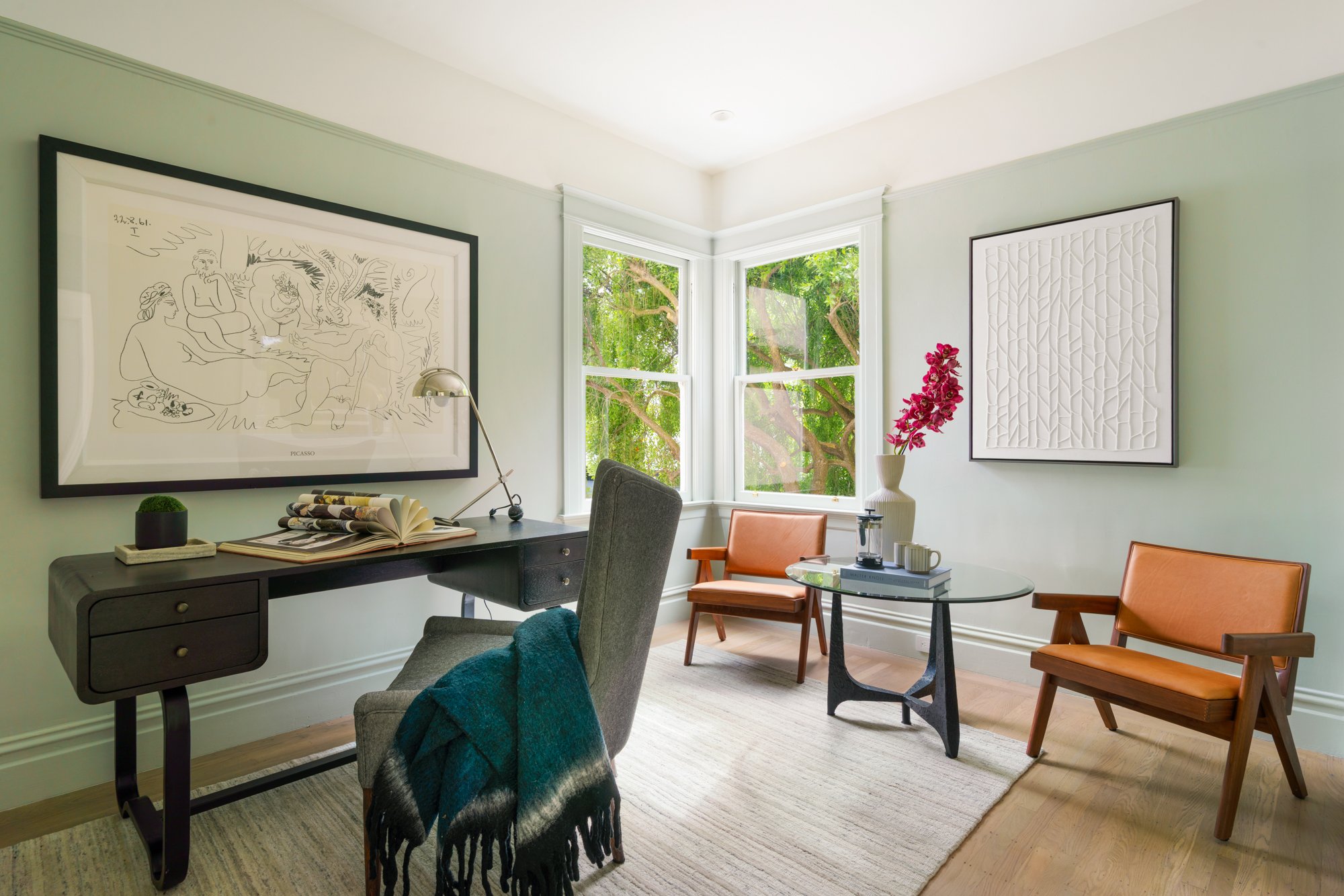Modern home office with a wooden desk, gray chair with blue throw, two leather chairs, a round glass coffee table with books and flowers, and two large framed artworks on light green walls.