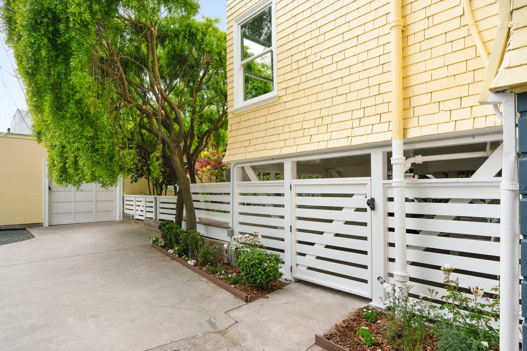Driveway beside a yellow house with white picket fence and trees